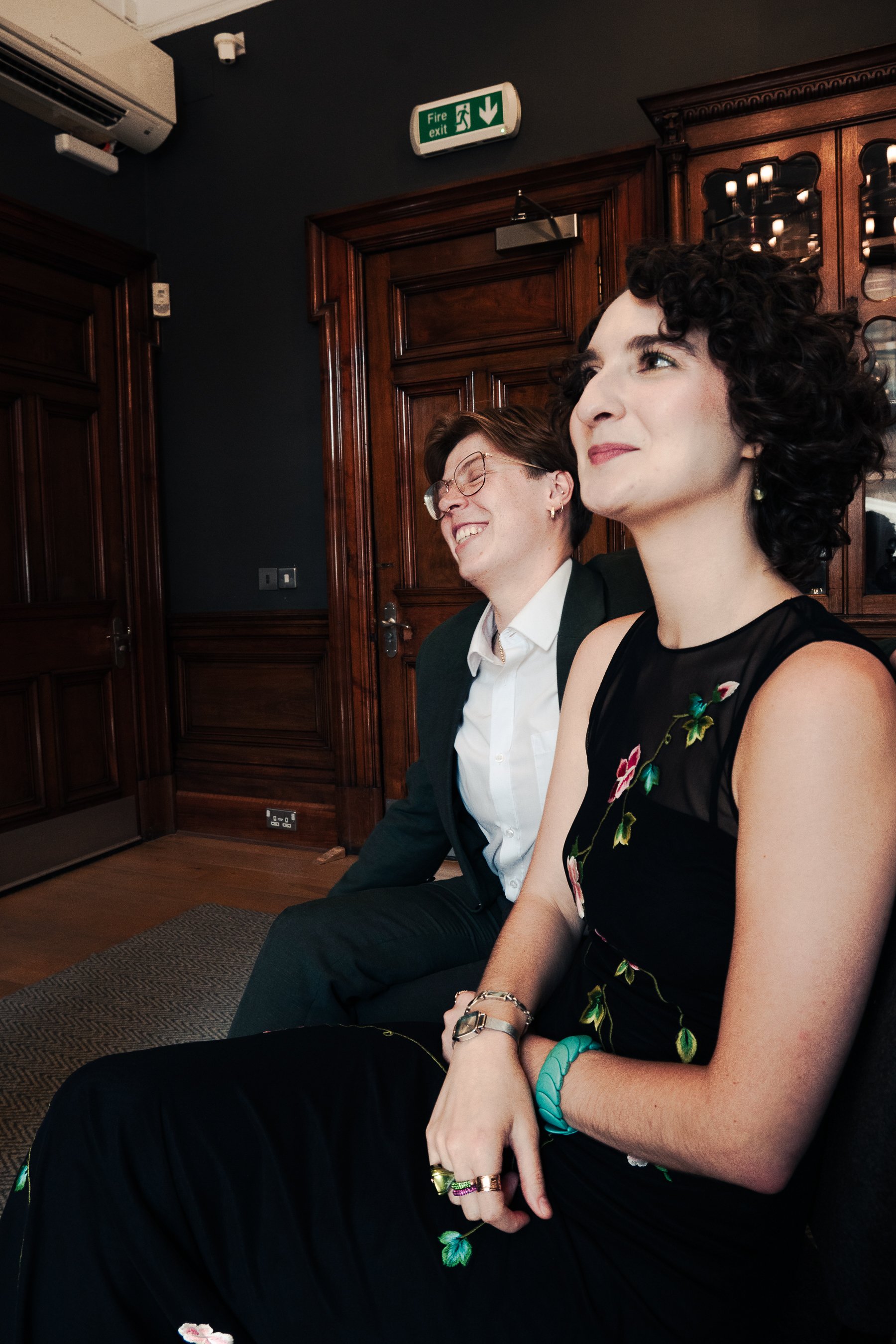 Two people sitting next to each other indoors, with wood-paneled walls and a fire exit sign above them. The woman in the foreground has curly dark hair, light skin, and is wearing a black dress with floral embroidery, bracelets, and rings. The man be