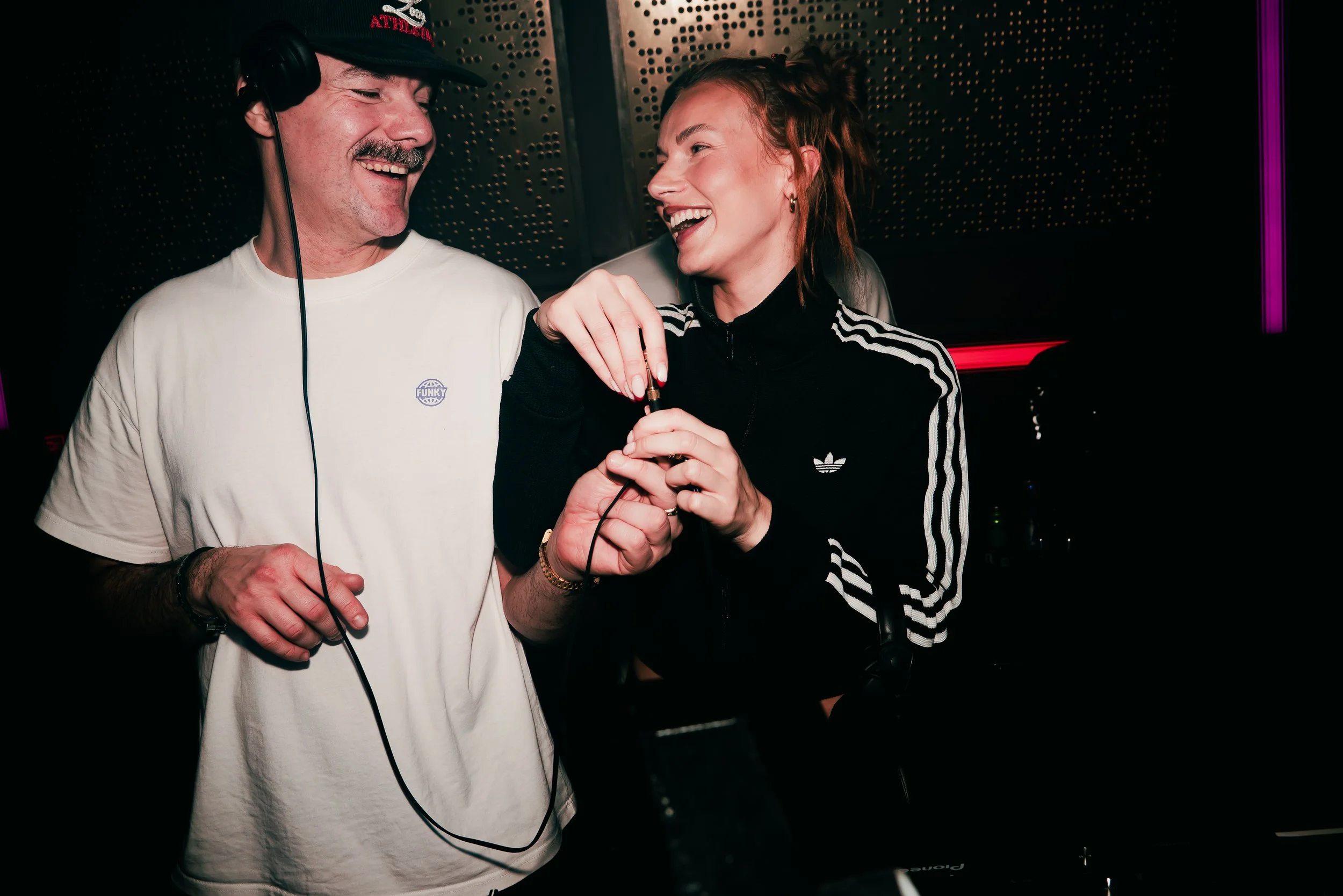 A man and a woman are smiling and talking to each other while standing close together, with the man wearing headphones and the woman holding a microphone. The background suggests they are in a nightclub or music venue.