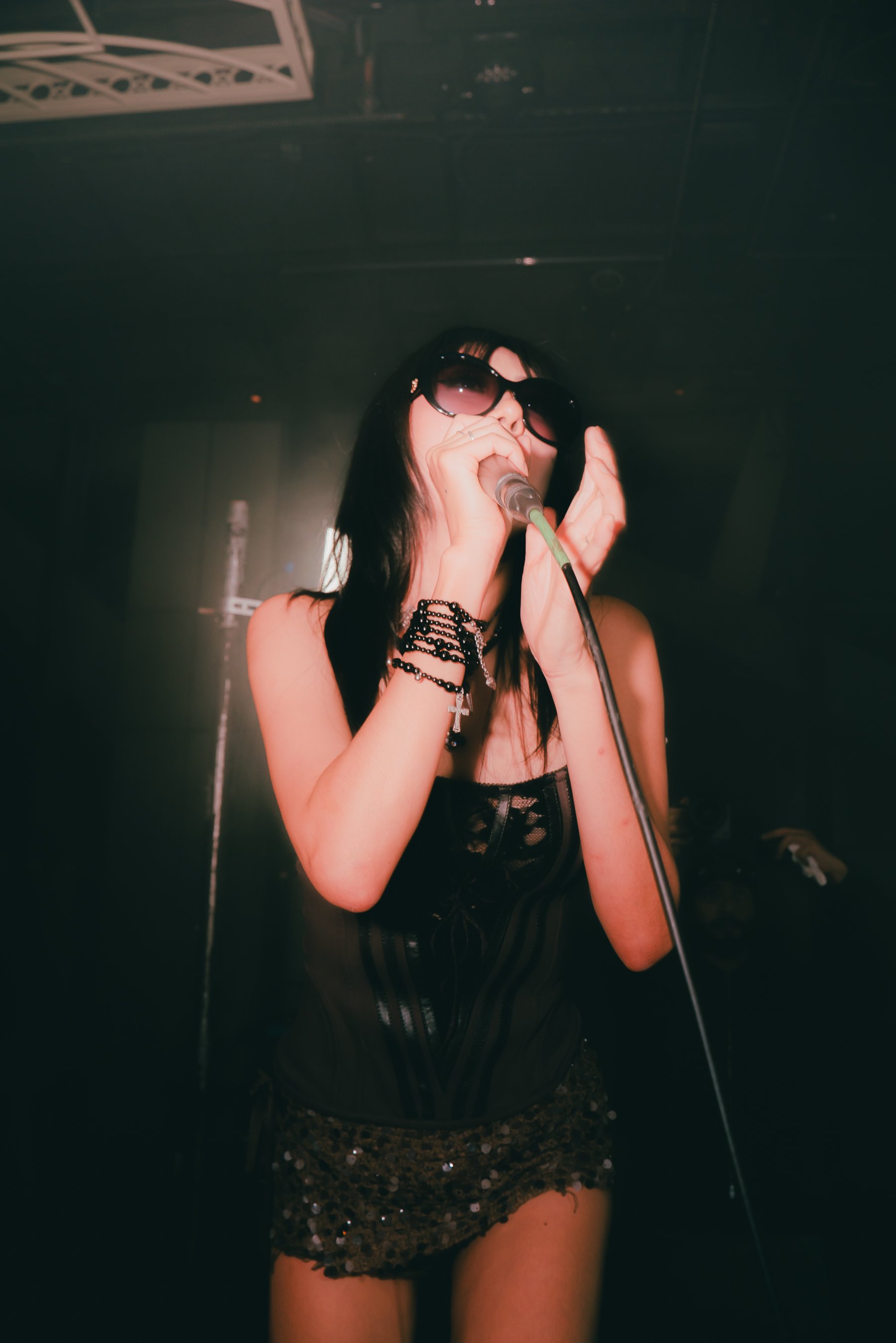 Woman singing into a microphone on stage, wearing large sunglasses, black lace top, and a sequined skirt.