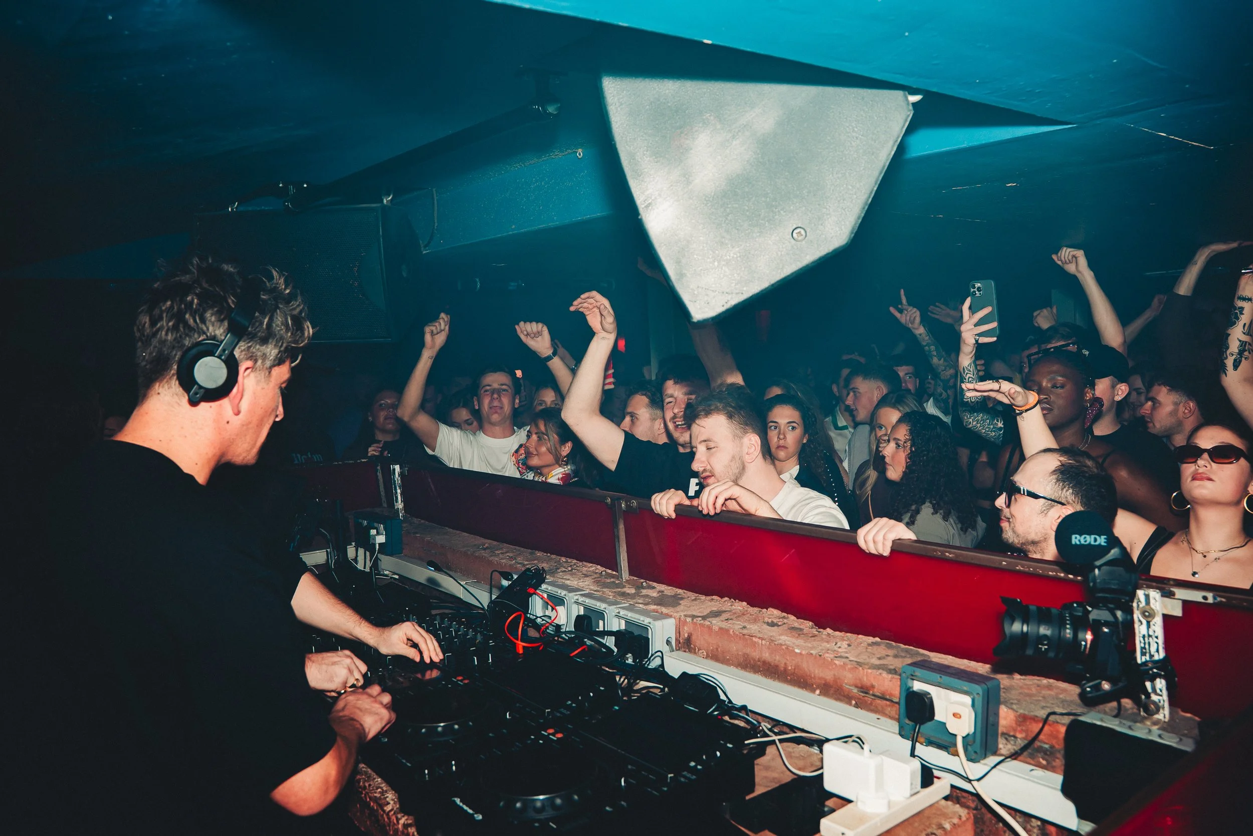 A DJ wearing headphones is performing at a crowded indoor music event. The crowd, diverse in appearance, is dancing and enjoying the music in front of the DJ booth, with some raising their hands and holding phones.