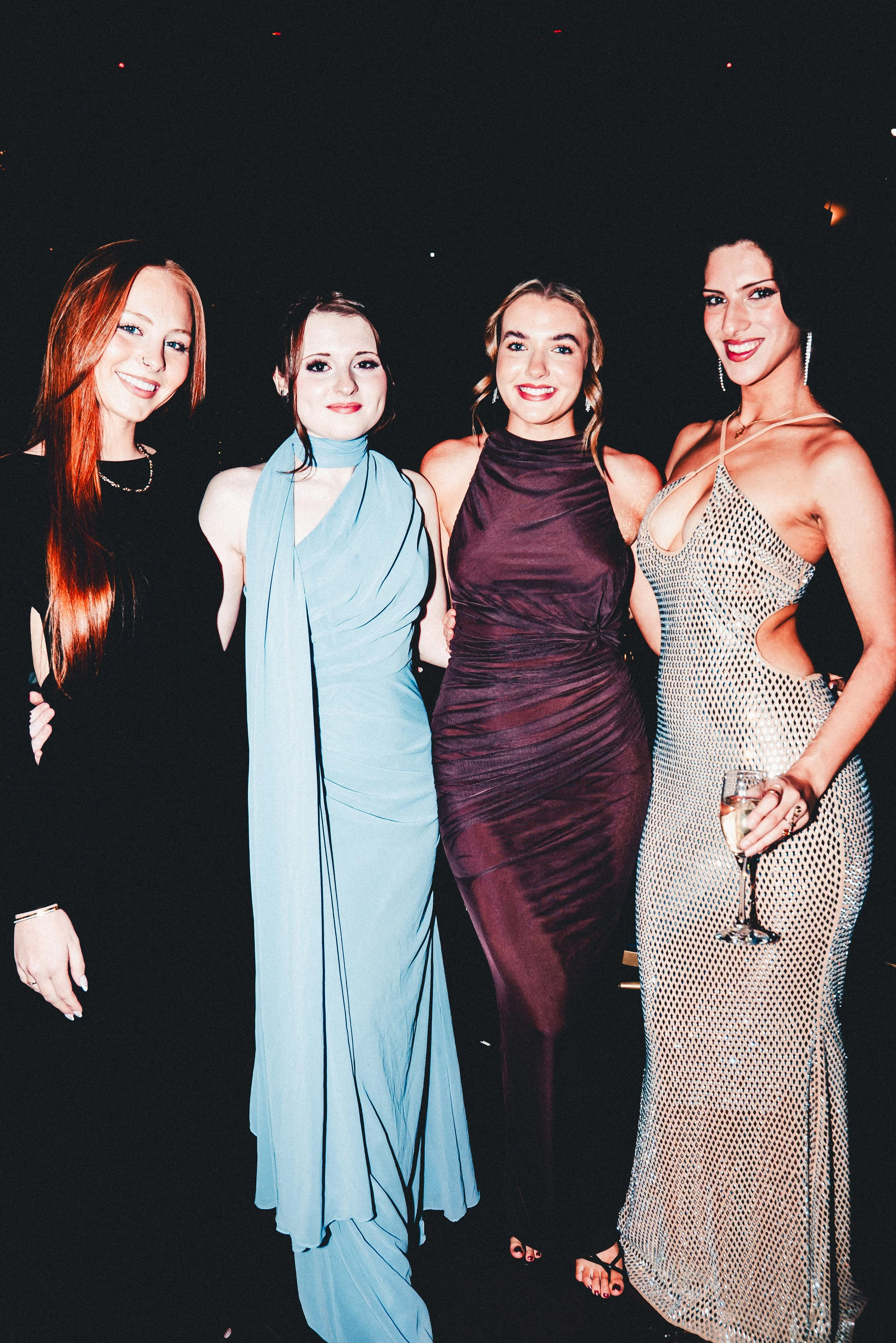 Four women in stylish evening dresses at a party, smiling and posing for the photo against a dark background.