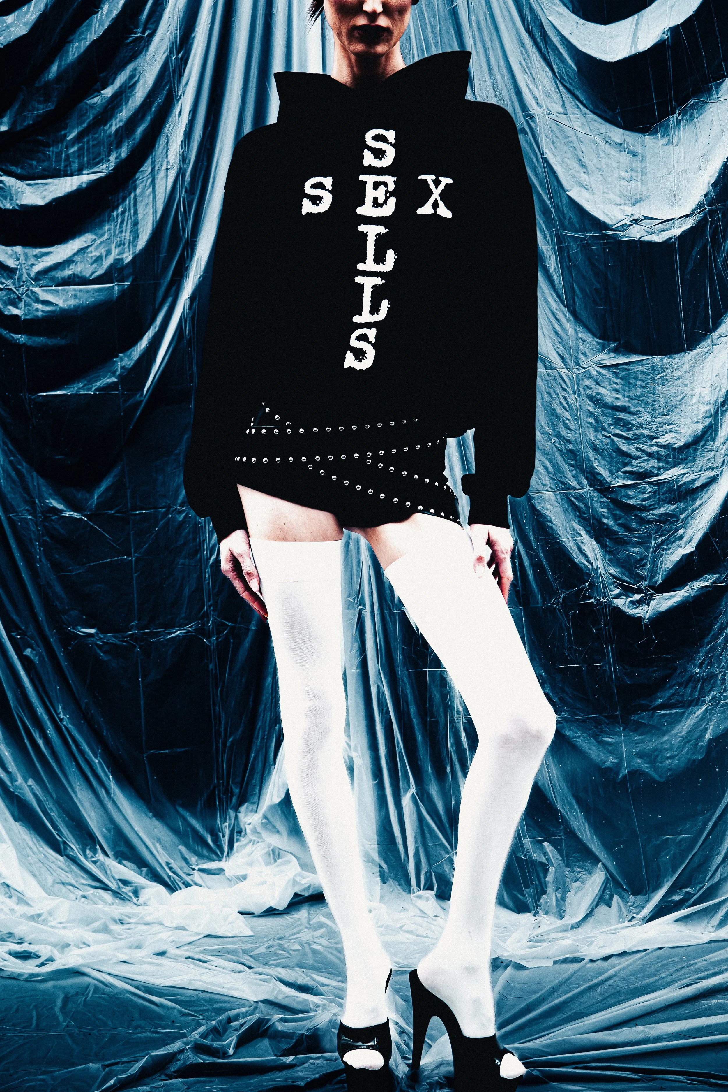 Person wearing a black hoodie with white text reading 'SEX SLIPS' on front, white thigh-high stockings, and black high heels, standing in front of silver metallic draped backdrop.