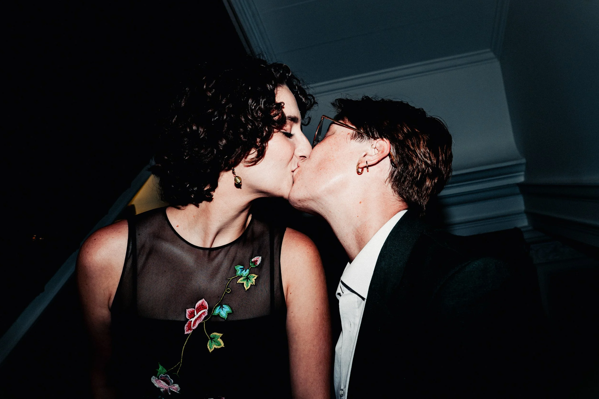 Two women sharing a kiss at a formal event. One woman has dark curly hair and is wearing a black dress with floral embroidery, and the other woman has short hair, glasses, and earrings, wearing a tuxedo.