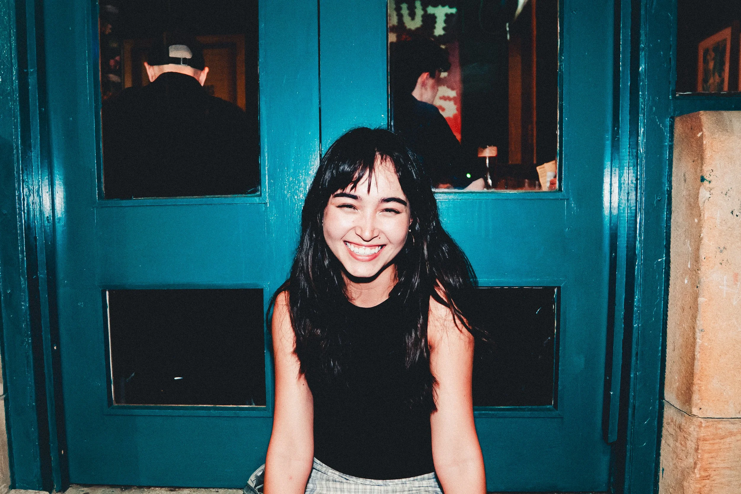 Young woman with long dark hair smiling while sitting in front of a blue door with two men in the background.