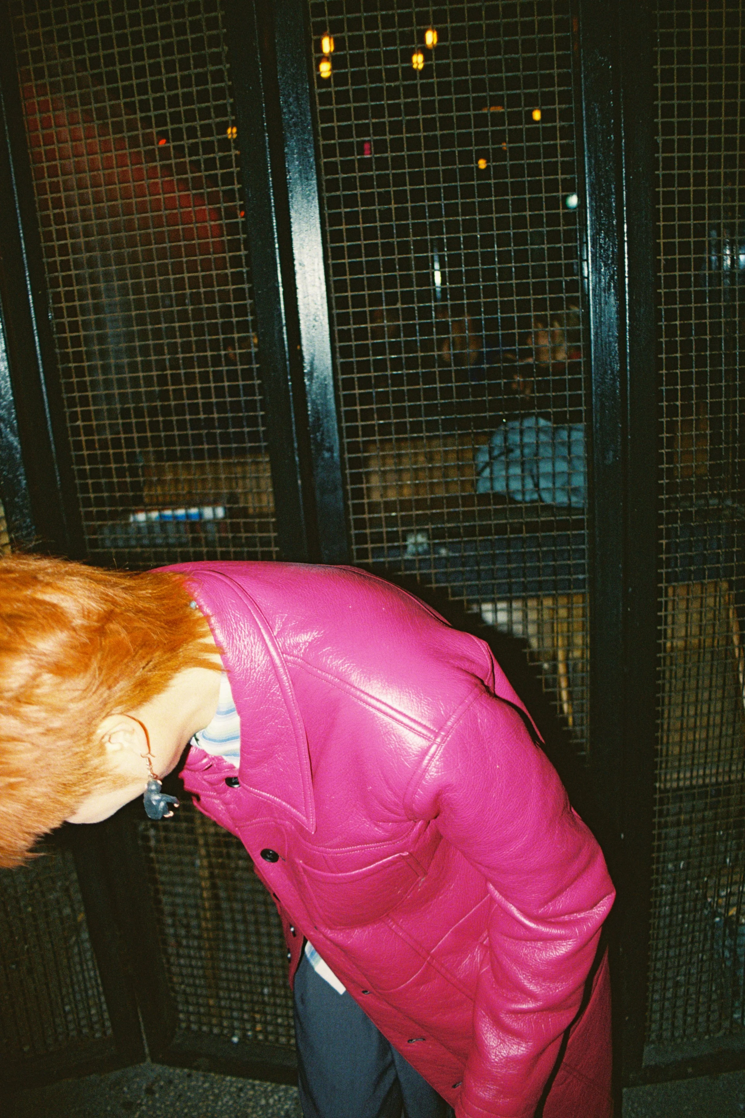 A person with reddish hair wearing a pink leather jacket, standing in front of a black metal gate or fence.