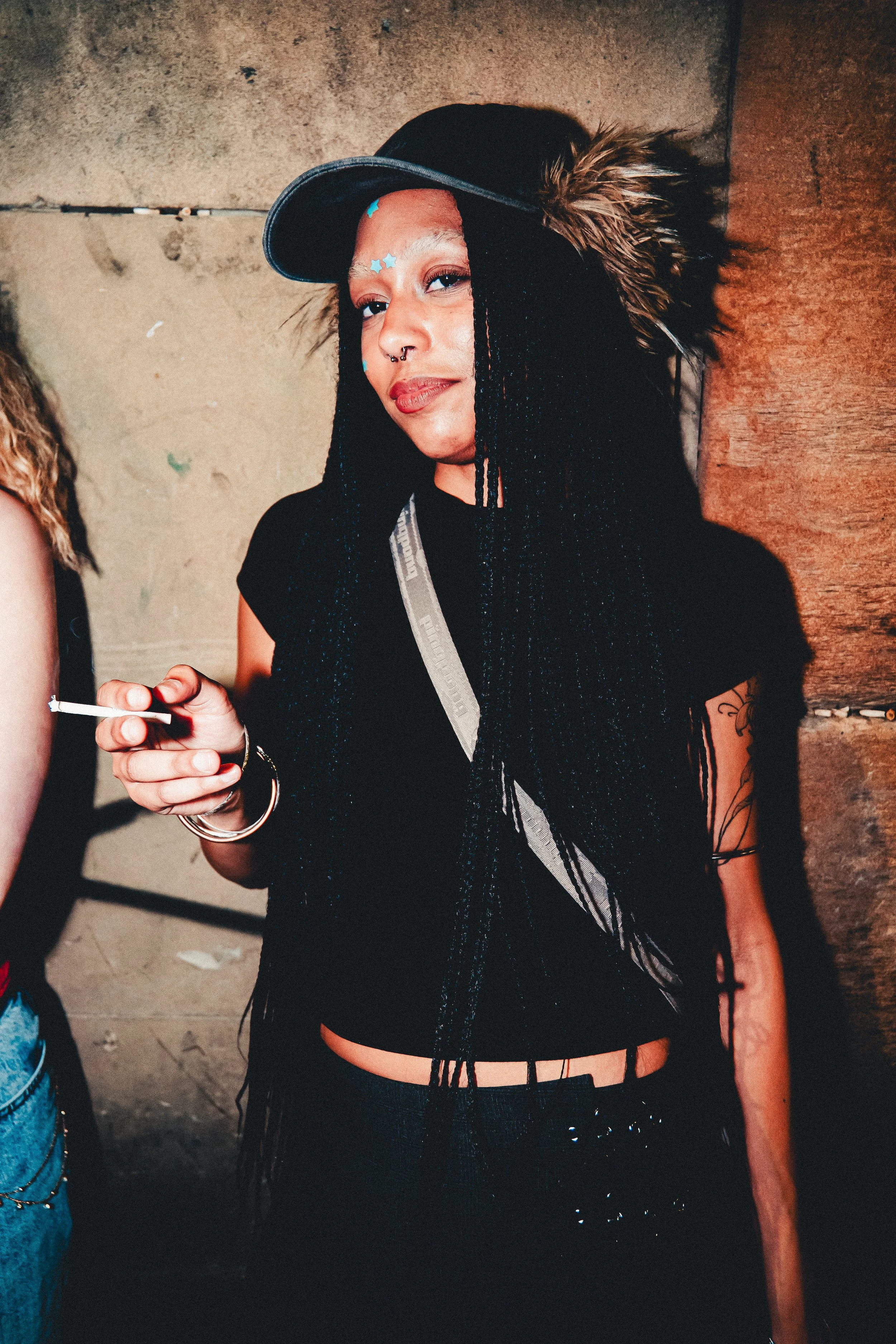 A woman with long black braided hair, wearing a black fringed top and a black baseball cap, stands against a textured wall. She has star-shaped stickers on her face, a septum piercing, and is holding a lit cigarette.