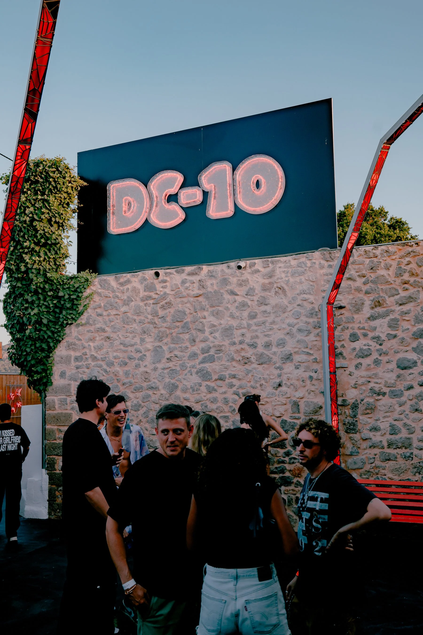 People gathered outdoors in front of a pink stone wall with a large sign that reads 'DC-10'. The group is engaged in conversation, with some smiling and others looking around. The sky is clear and blue, and there are trees and metal structures visibl