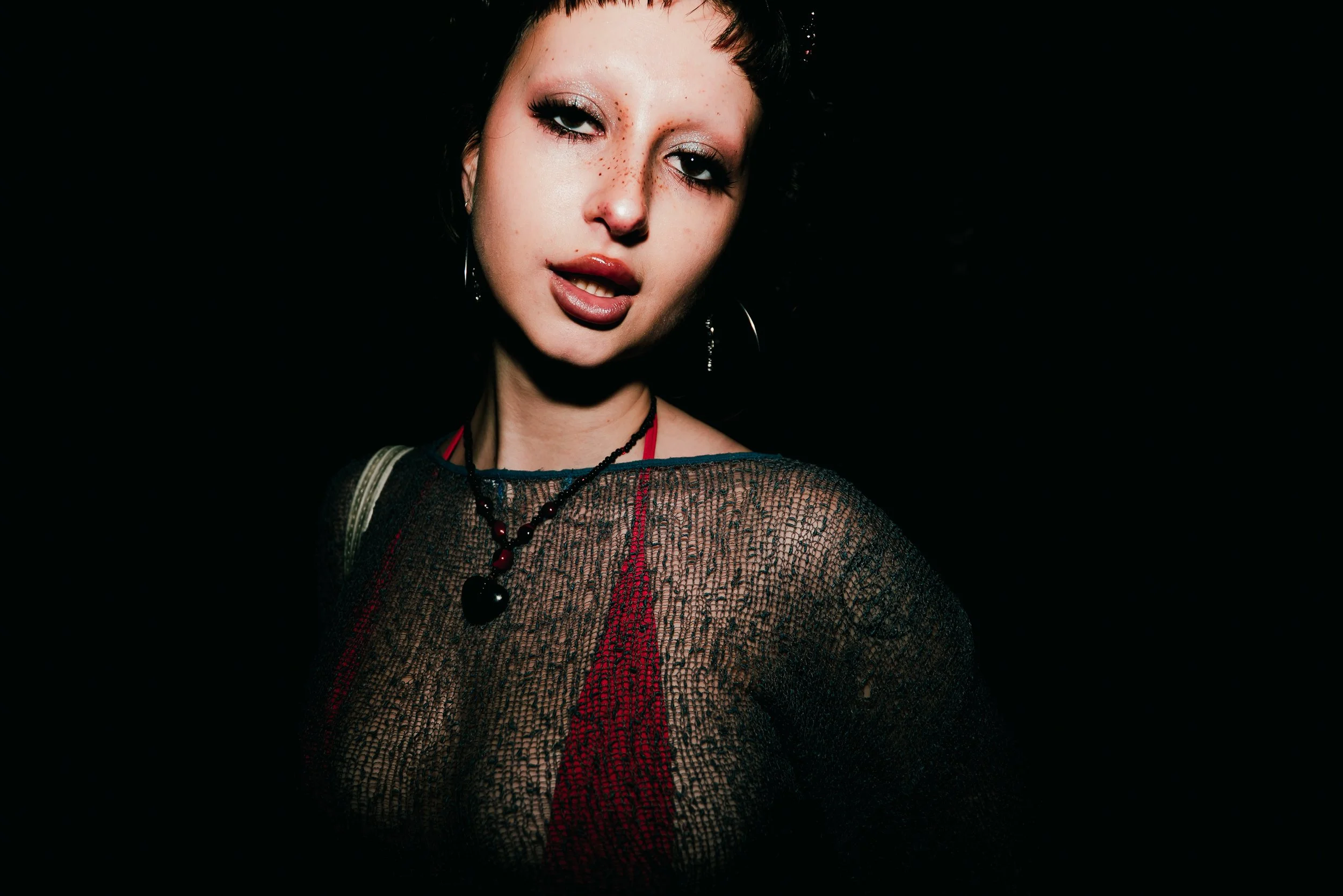 A woman with short dark hair and makeup with glitter on her eyelids and freckles on her face, wearing a black mesh top, a red lace dress, and a necklace with a heart pendant against a black background.