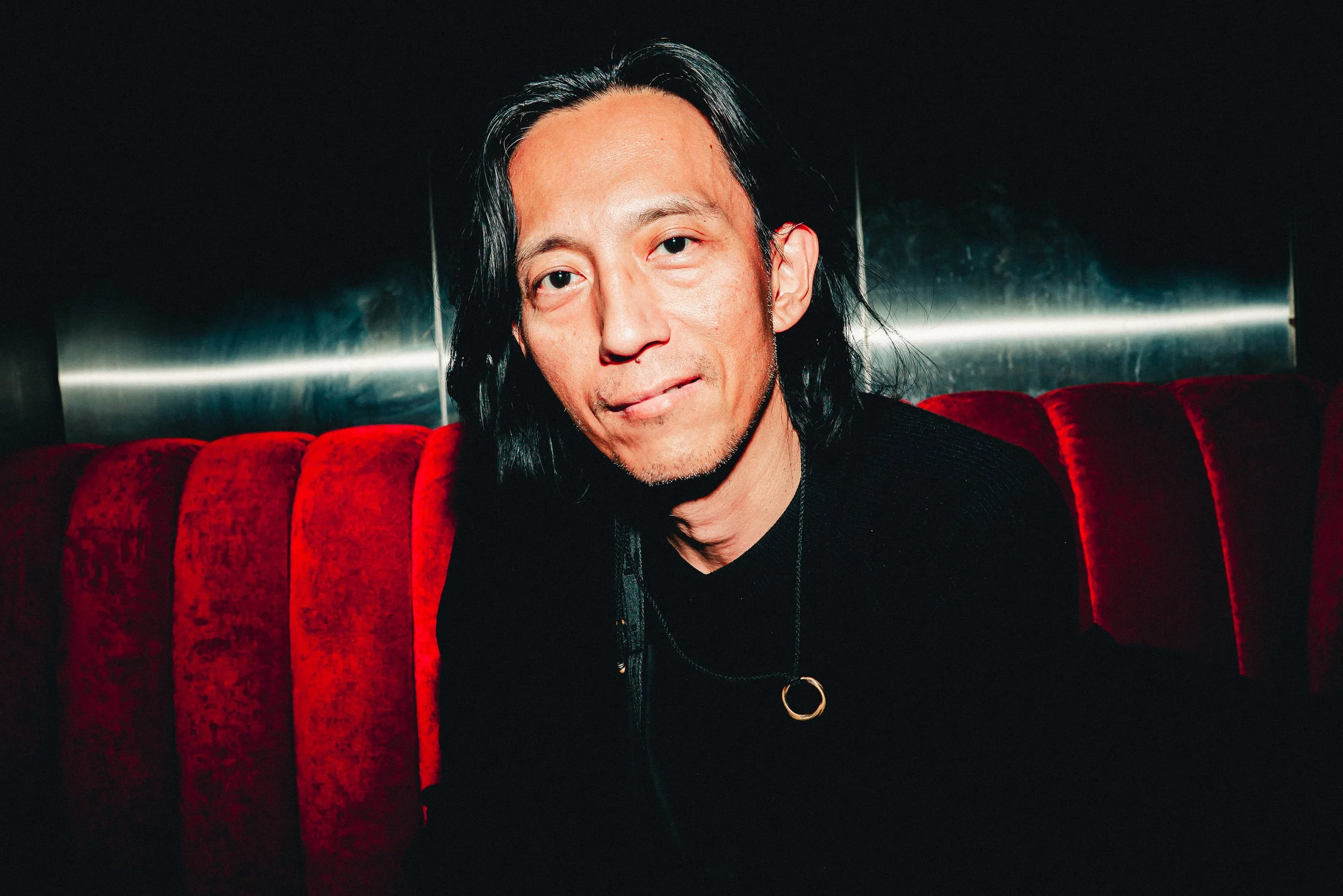 Portrait of a man with long black hair, light skin, and wearing a black shirt with a gold ring necklace, sitting on a red velvet sofa against a dark background.