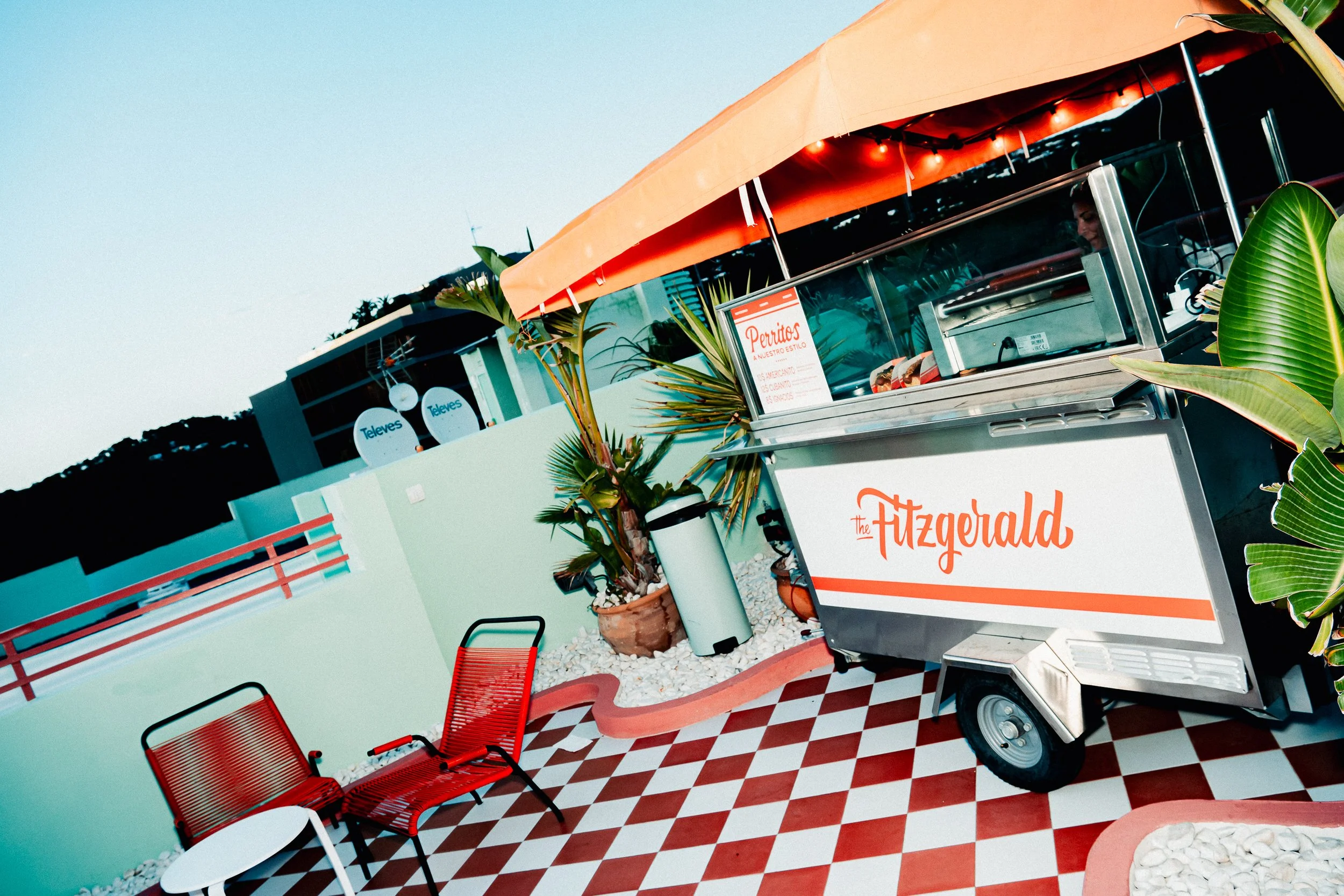 Colorful retro-style food stand with a sign that says 'The Fitzgerald' surrounded by red chairs and potted plants, with a checkered red and white floor.