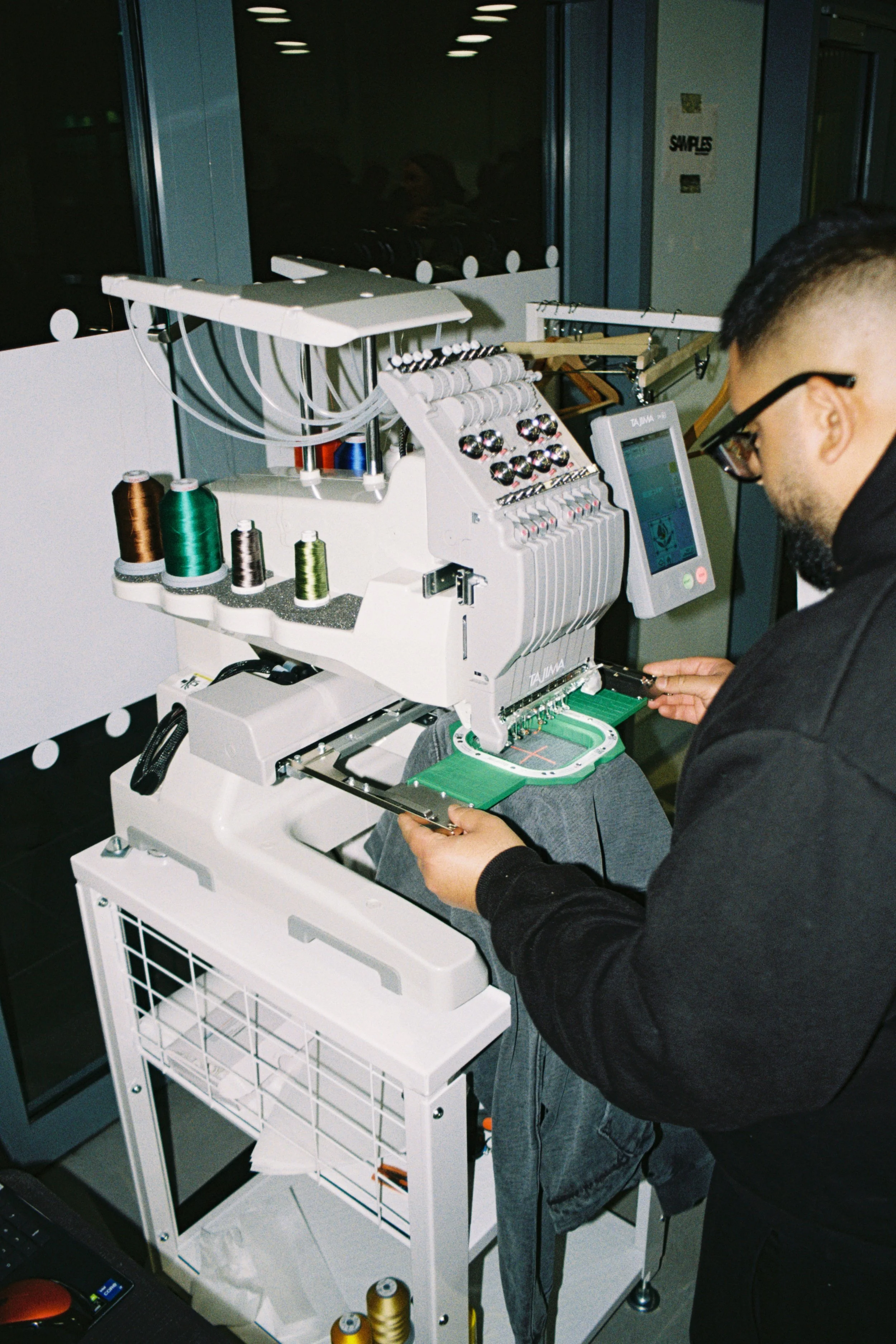 Person operating a sewing machine in an indoor setting, with spools of thread on a white cart nearby and work lights above.