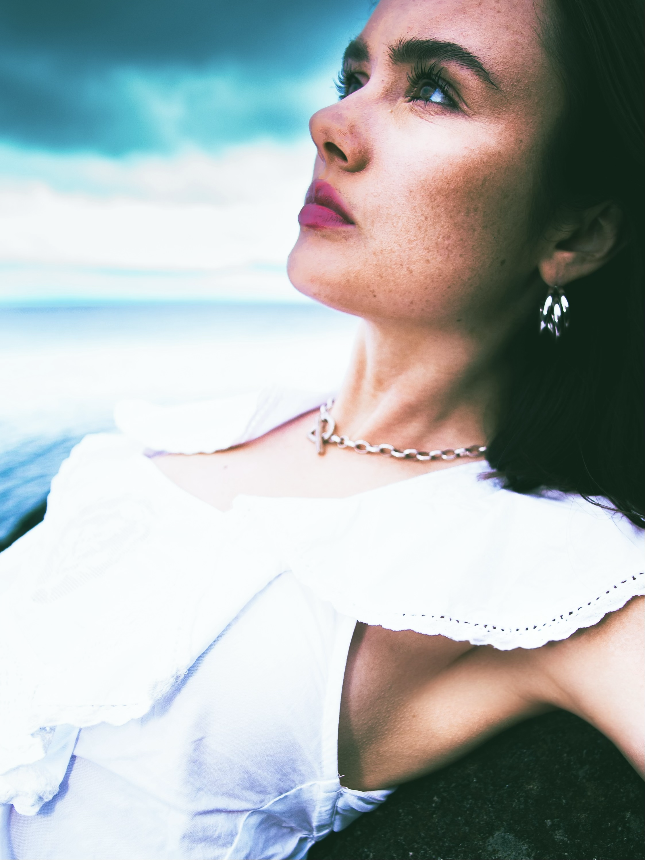 Close-up of a woman with dark hair and freckles, looking upwards and to the left, with a cloudy sky and ocean in the background. She is wearing earrings, a necklace, and a white off-shoulder top.
