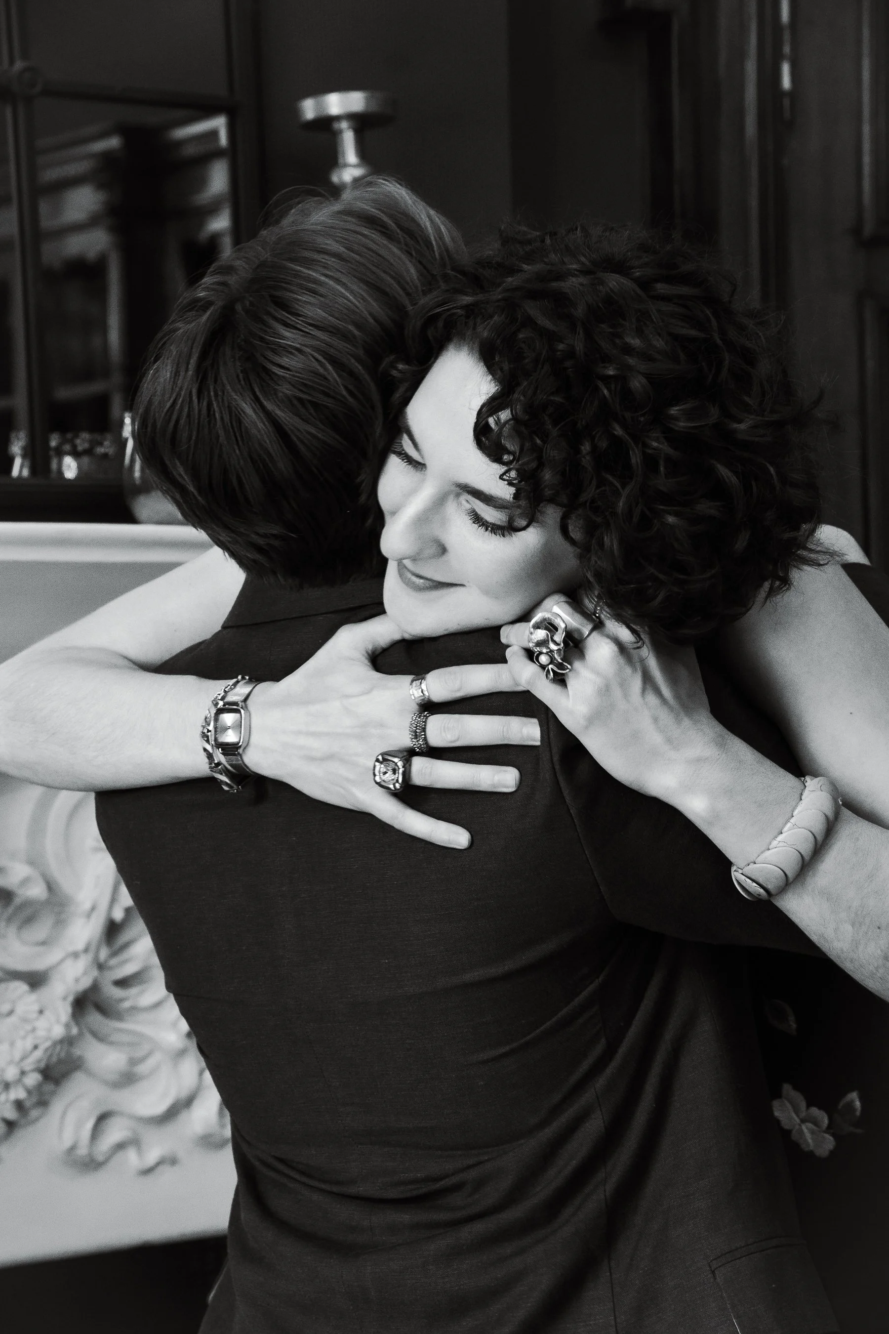Two people embracing warmly, with one person's face visible and the other person's face hidden; they are wearing jewelry and are in an indoor setting with dark furniture in the background.