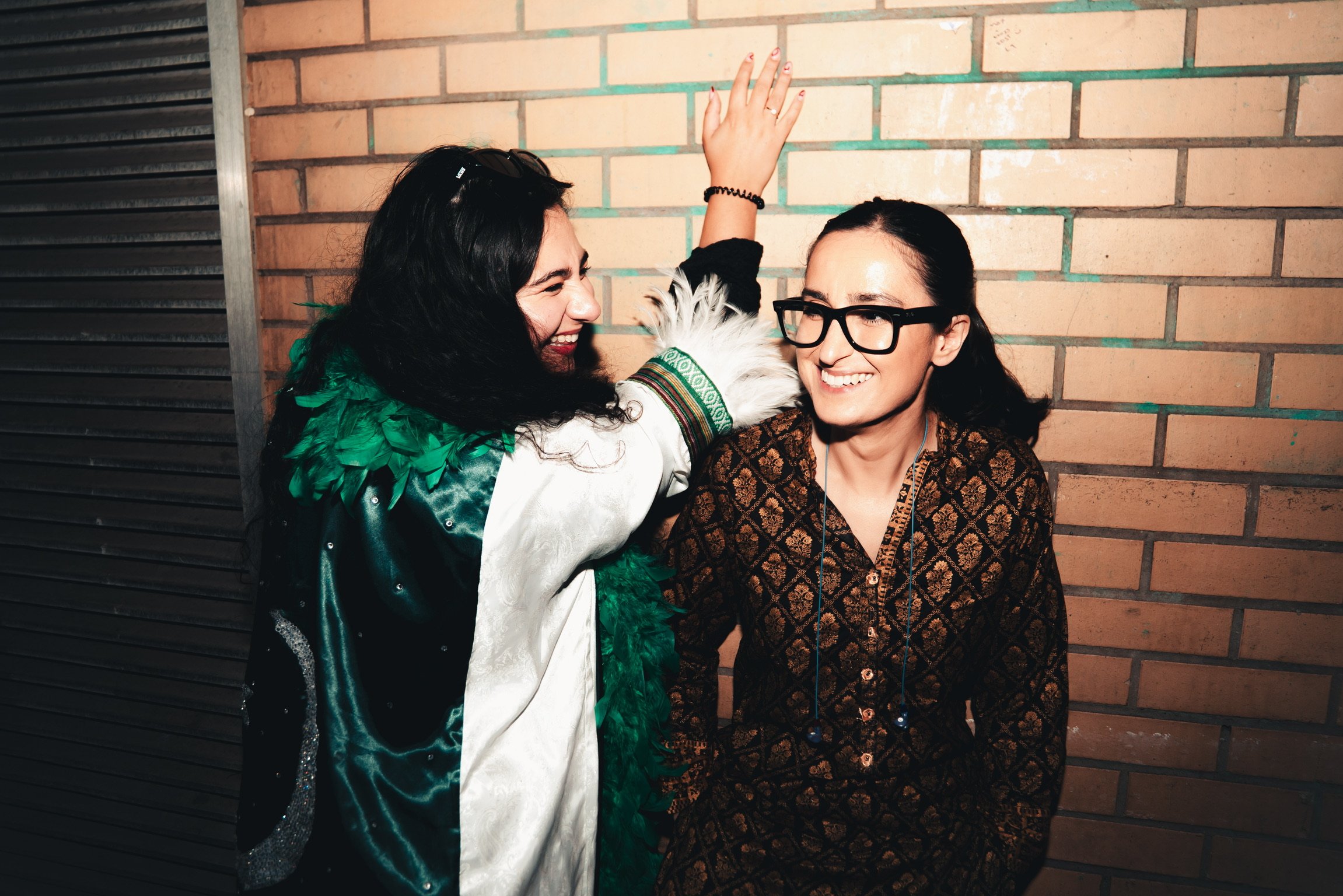 Two women smiling and laughing together against a brick wall. One woman with dark hair, wearing glasses and a patterned shirt, and the other woman with dark hair, wearing sunglasses on her head and a jacket with green feathered detail, is raising her