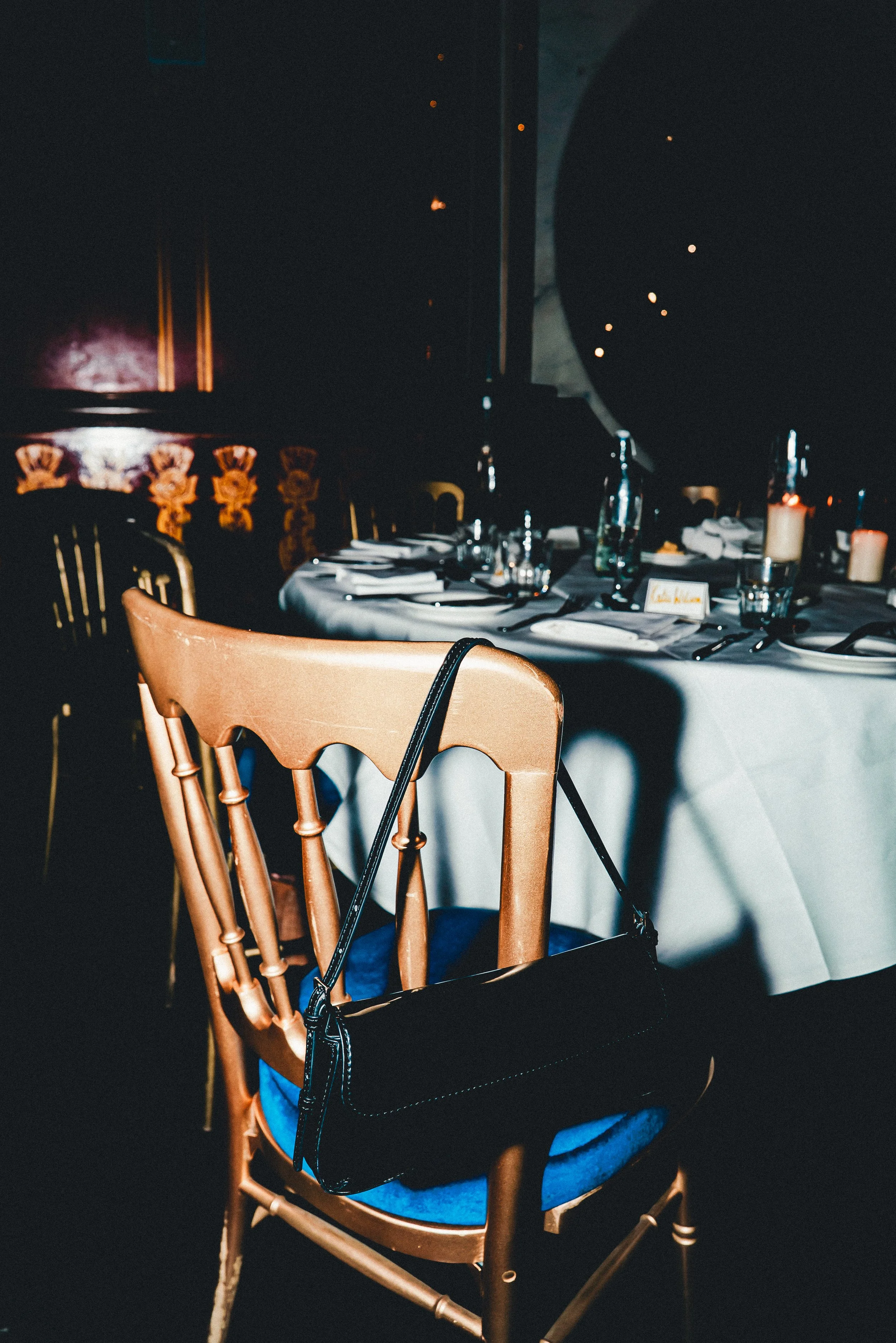A dining table set with plates, glasses, and candles in a dimly lit restaurant, with a beige wooden chair in the foreground.
