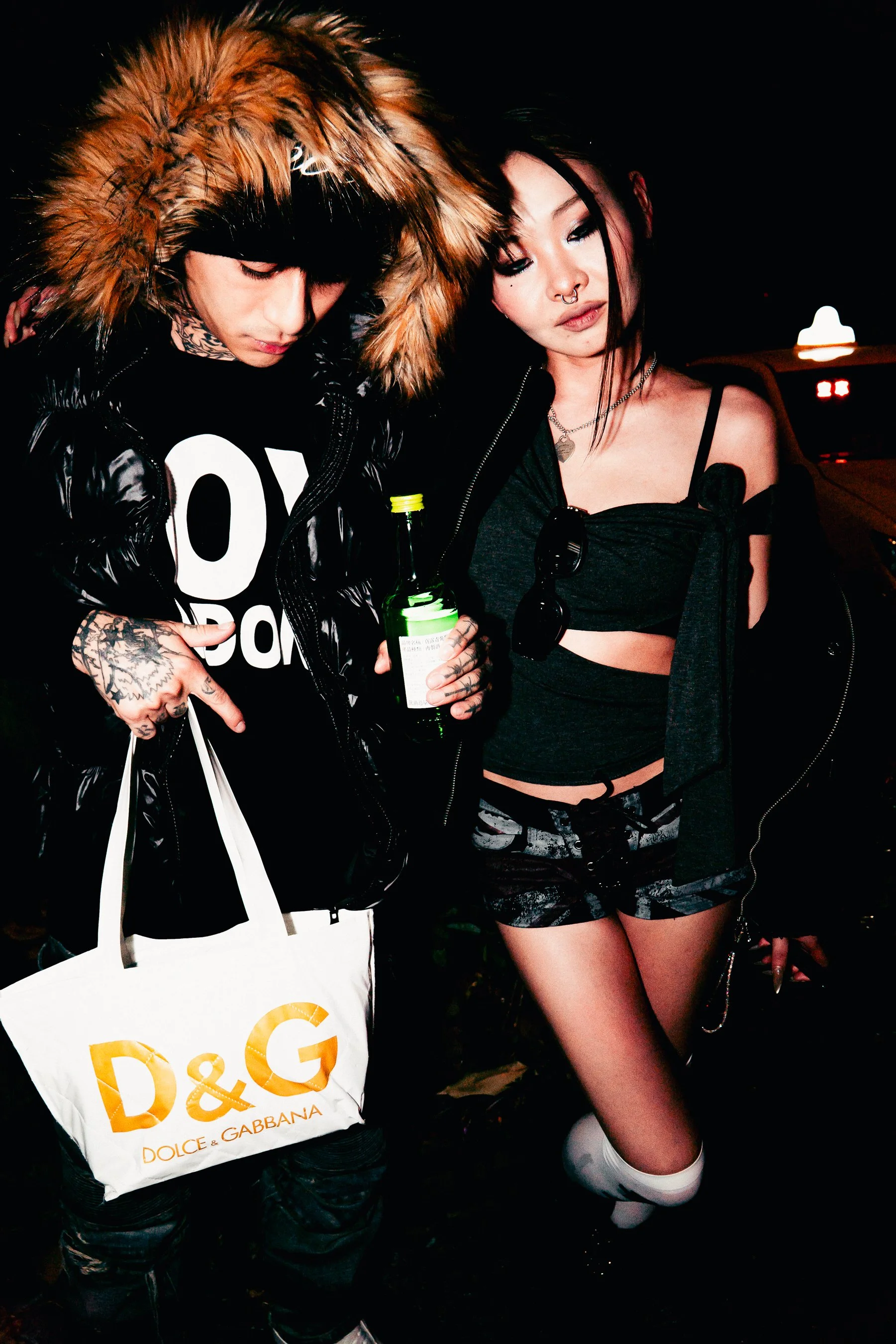 People at a party or nightclub, one person wearing a furry hooded coat and holding a D&G shopping bag, another person dressed in a black crop top, patterned shorts, thigh-high socks, and sunglasses hanging from her top.
