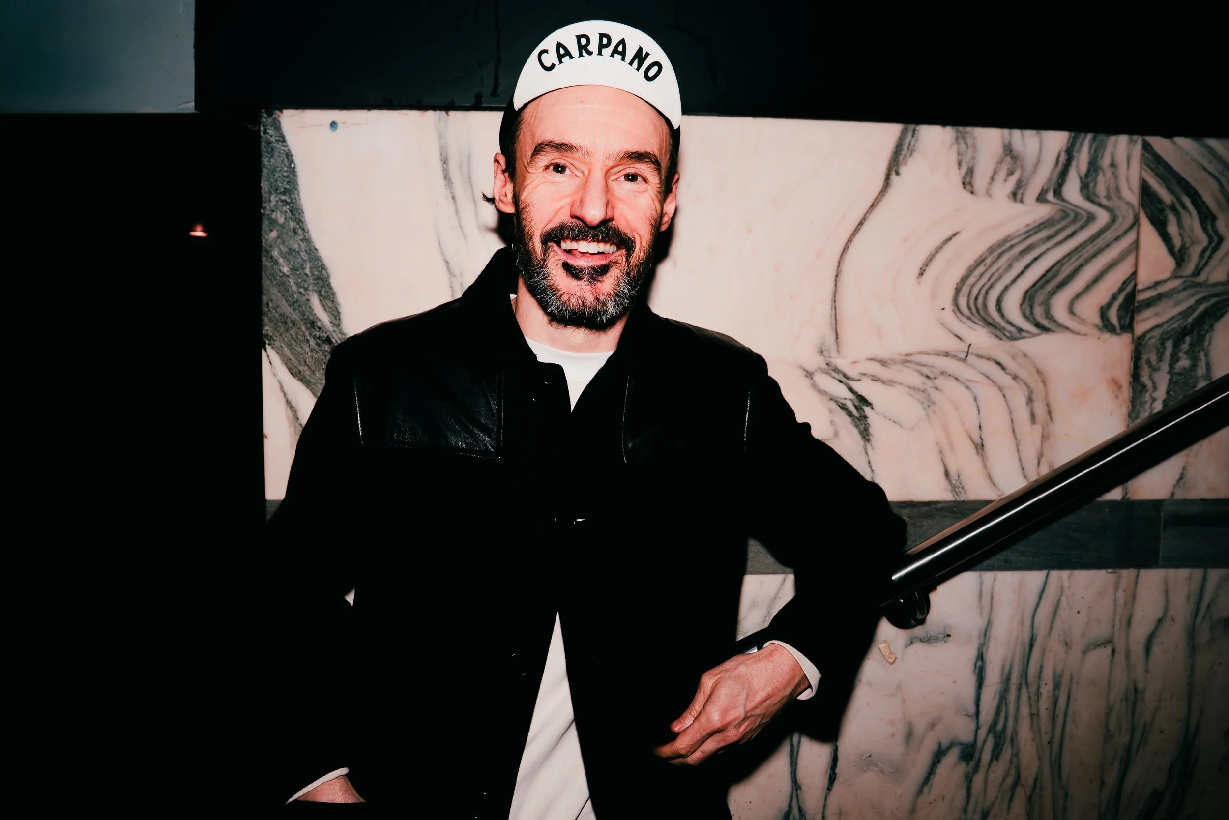 A man with a beard wearing a white cap with the name 'CARPANO' on it, smiling and dressed in a black and white outfit, standing in front of a marble wall.