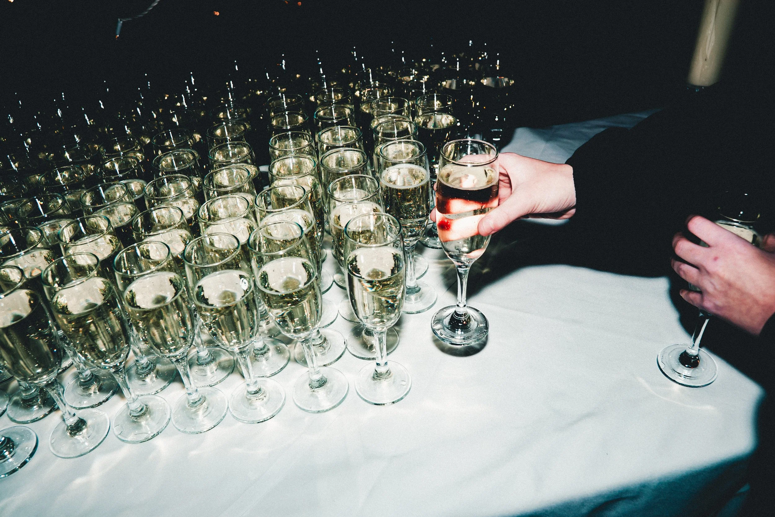 A person is pouring champagne into a tall glass among many filled champagne glasses on a table.