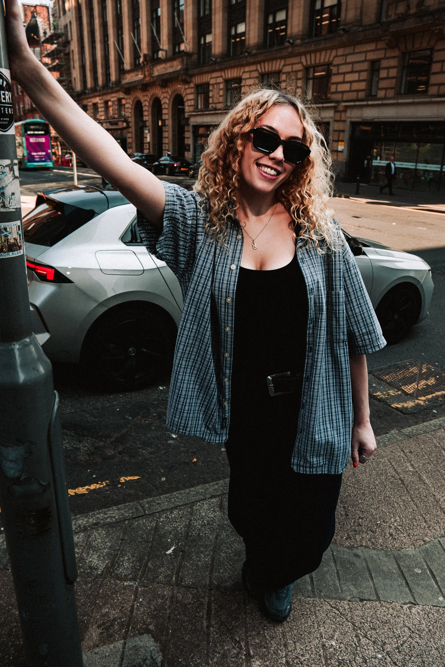 A smiling woman with curly blonde hair, wearing black sunglasses, a black top, and a plaid short-sleeved shirt, standing on a city sidewalk near a parked silver car and a lamppost with stickers, with city buildings and a bus in the background.