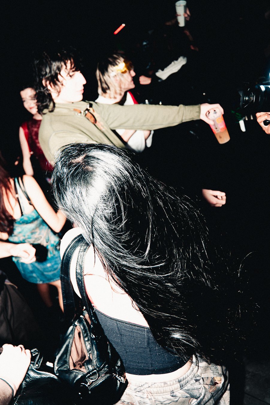 People dancing and socializing at a party or nightclub, with one person reaching out with a drink in hand and others holding drinks, in a dark environment with colorful lighting.