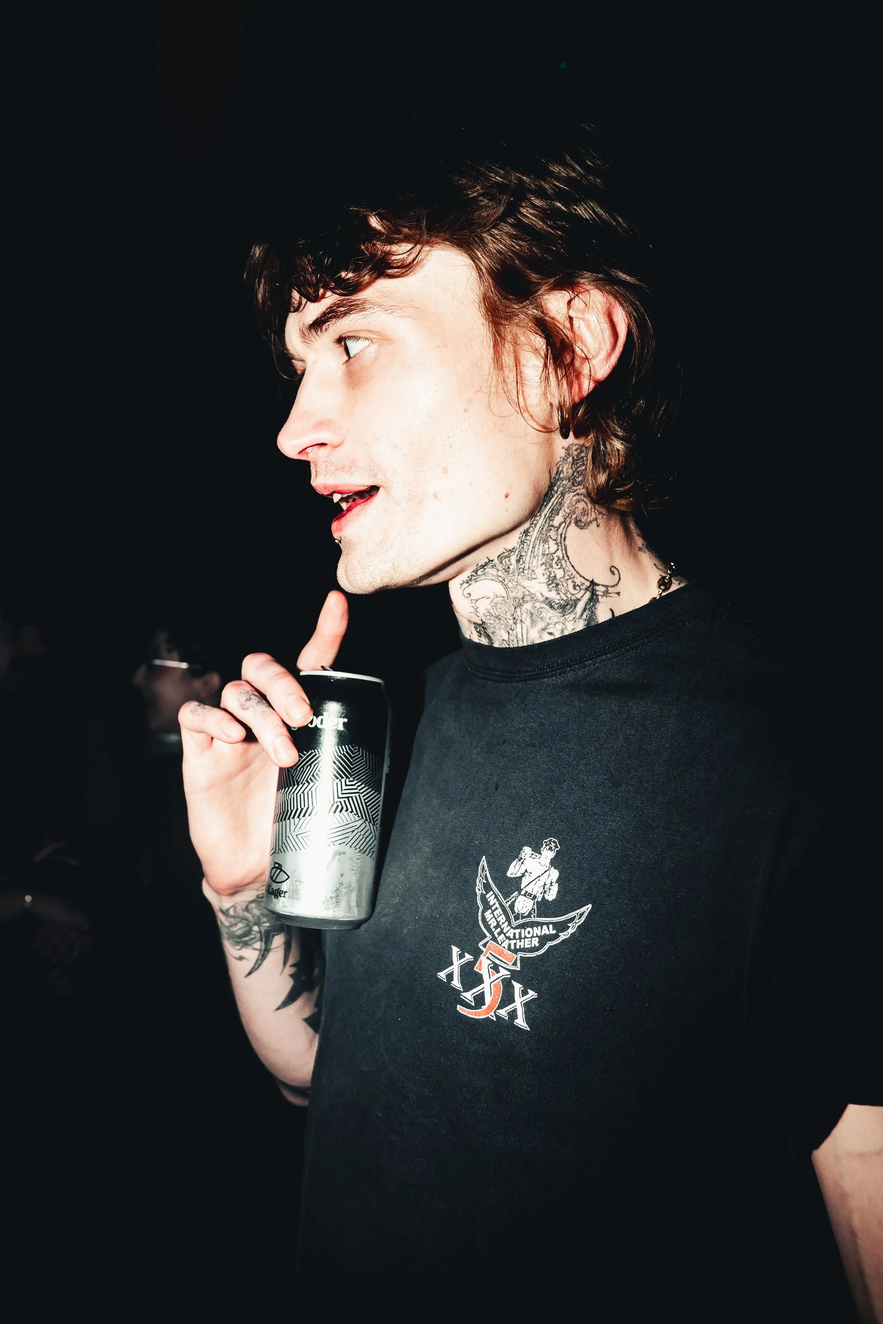 A young man with tattoos on his neck and arms, short dark hair, holding a silver can, wearing a black T-shirt with a graphic design.