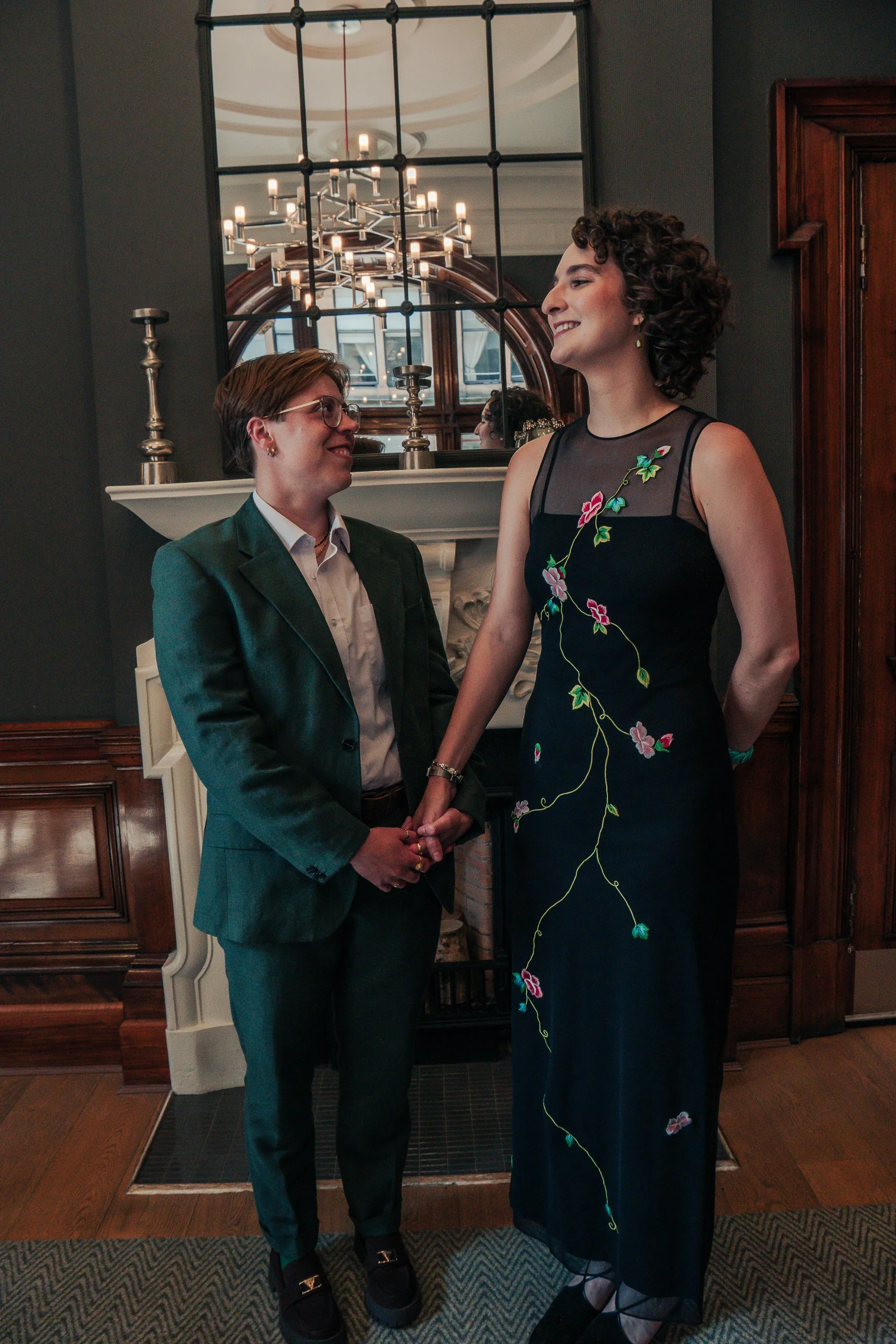 Two women holding hands, standing in a living room with a fireplace, mirror, and chandelier, smiling at each other.