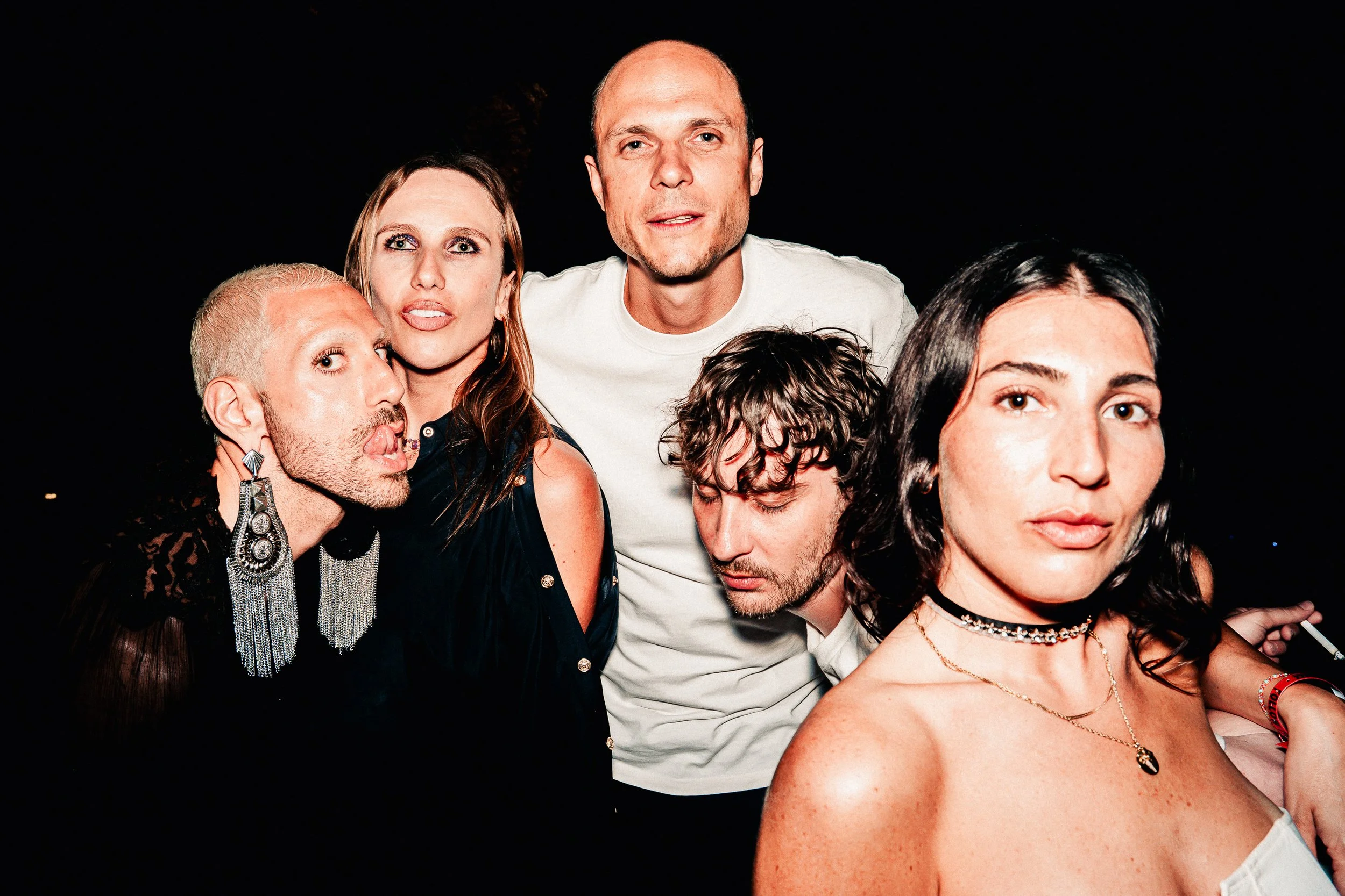 Group of six diverse friends smiling and posing closely together against a black background, with three women and three men, dressed in casual and trendy clothing, some wearing accessories.