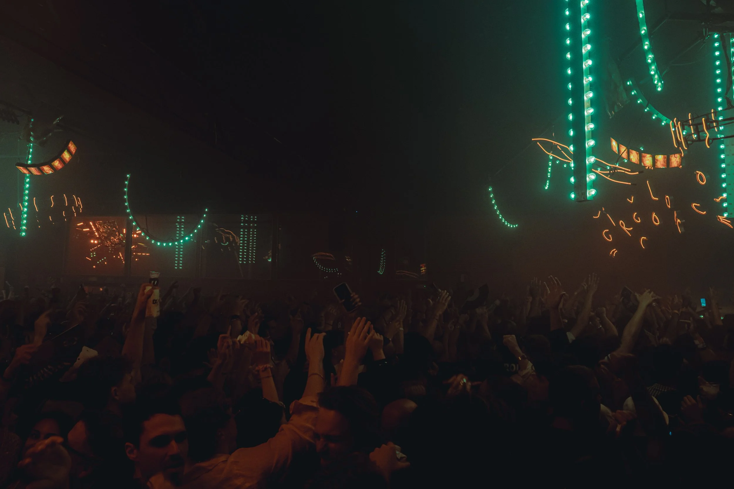 Crowd at concert or music event with raised hands and green and orange neon lights overhead.