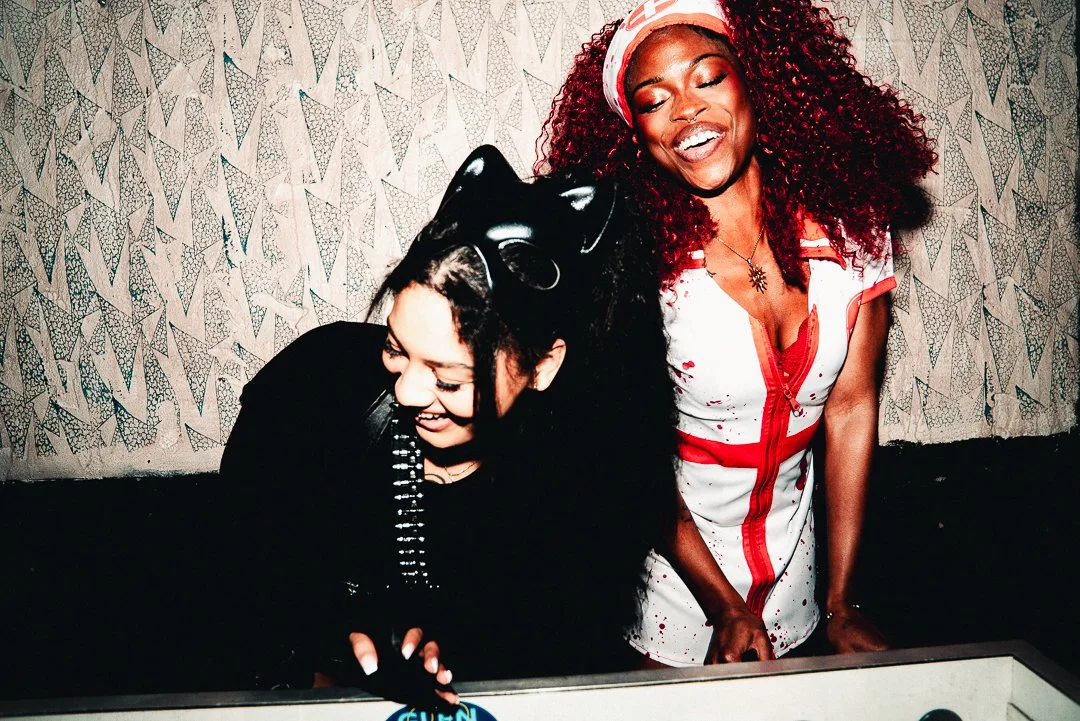 Two women with dark skin, one with curly red hair wearing a nurse costume with blood splatters and the other with dark hair and cat ears, laughing together