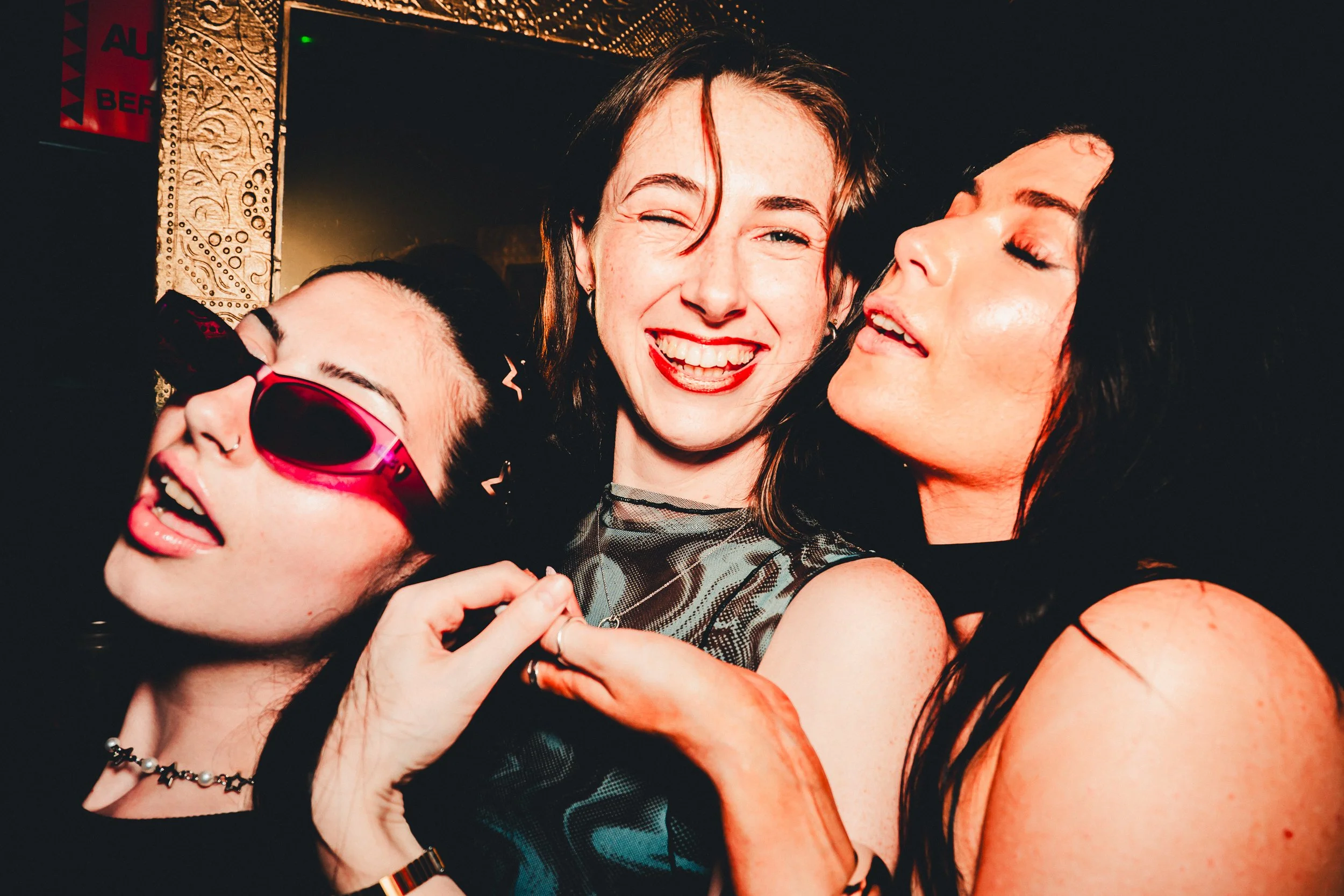 Three women happily dancing and hugging each other in a lively, colorful party setting.
