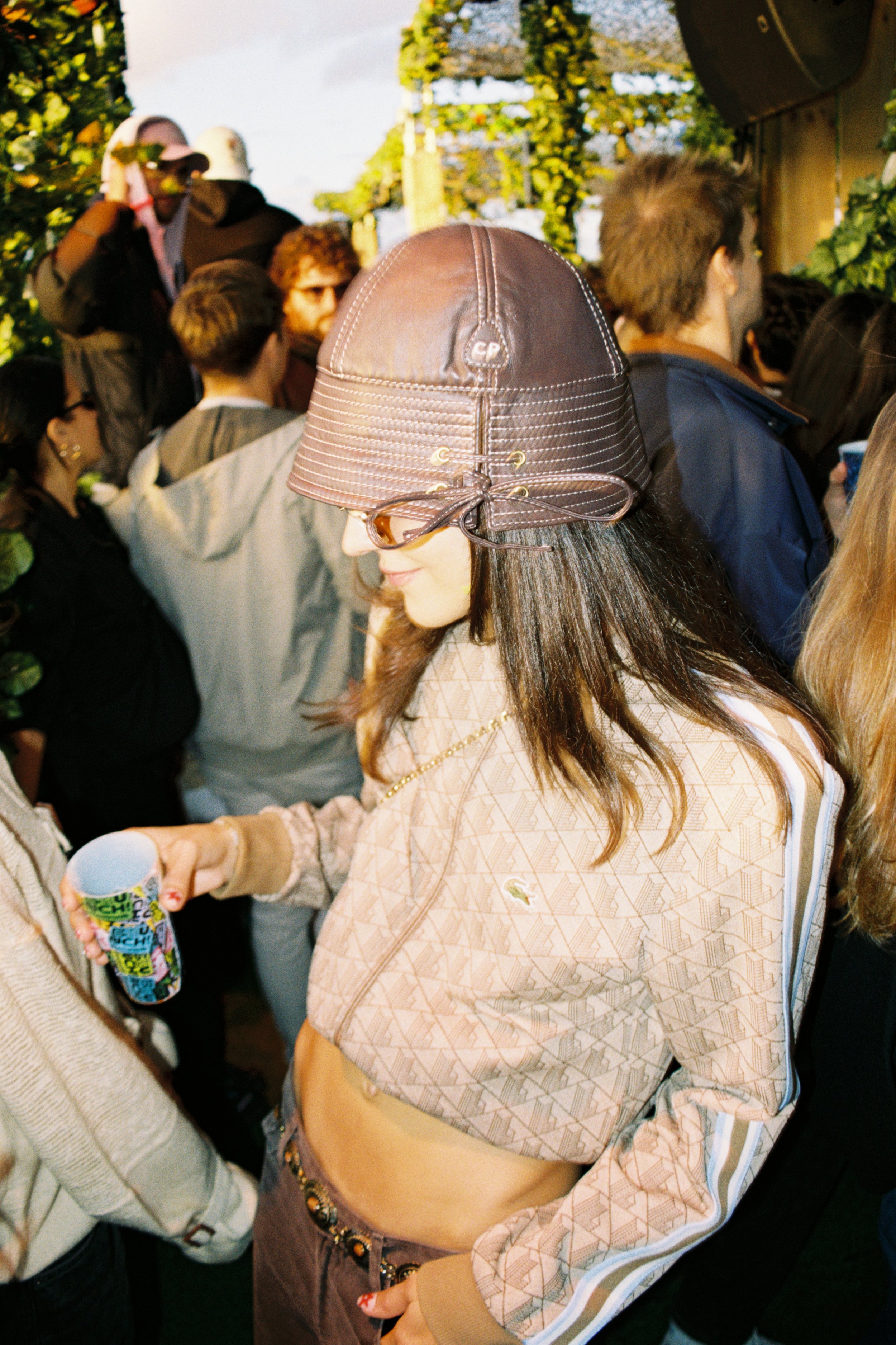 Close-up of a young woman wearing a large brown leather hat, patterned long-sleeve shirt, and beaded belt, holding a colorful cup, in a crowd outdoors.
