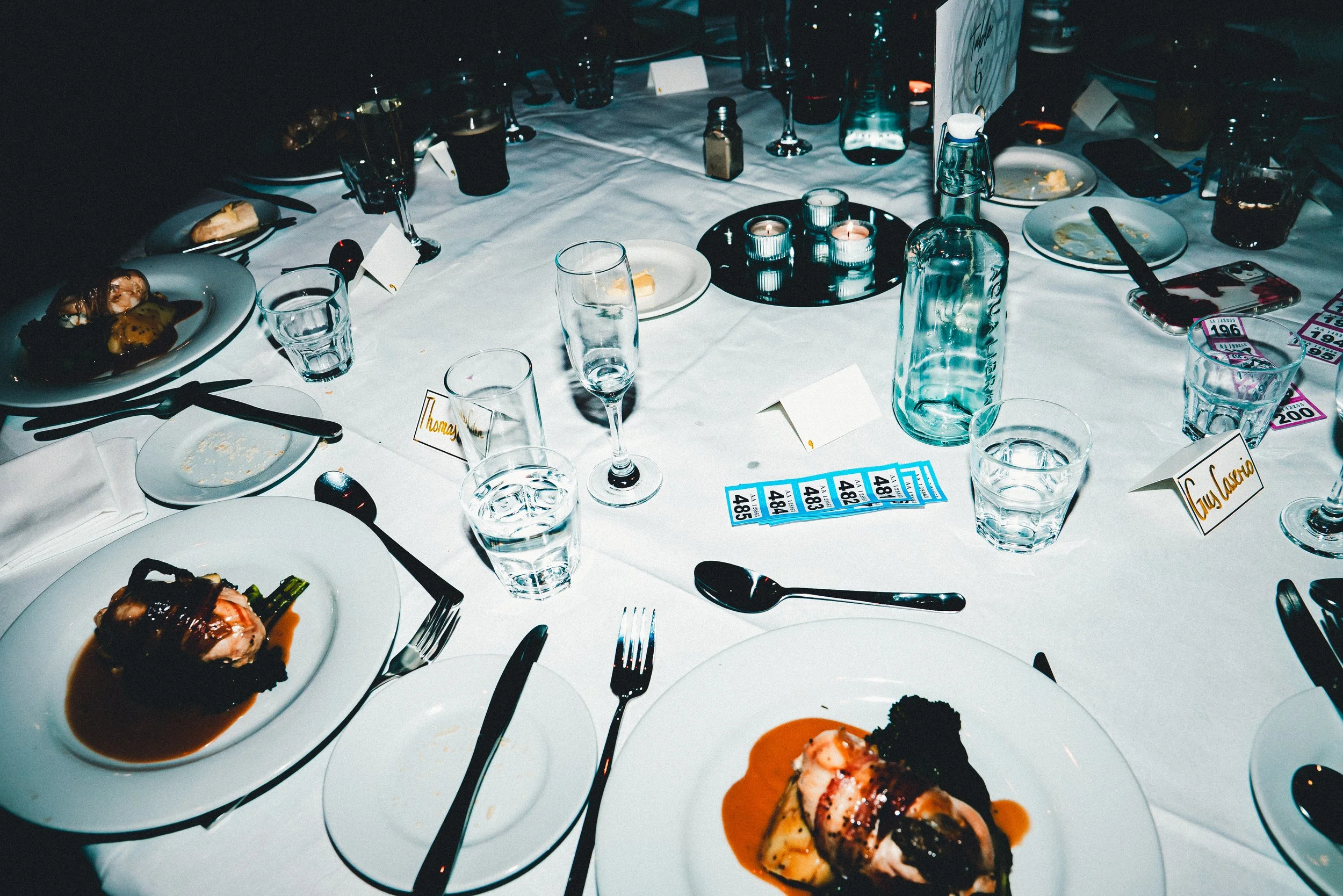 A table set for a meal with empty plates, glasses, utensils, and some partially eaten dishes, including meat with sauce, candles, a water bottle, and name cards at an elegant dinner event.