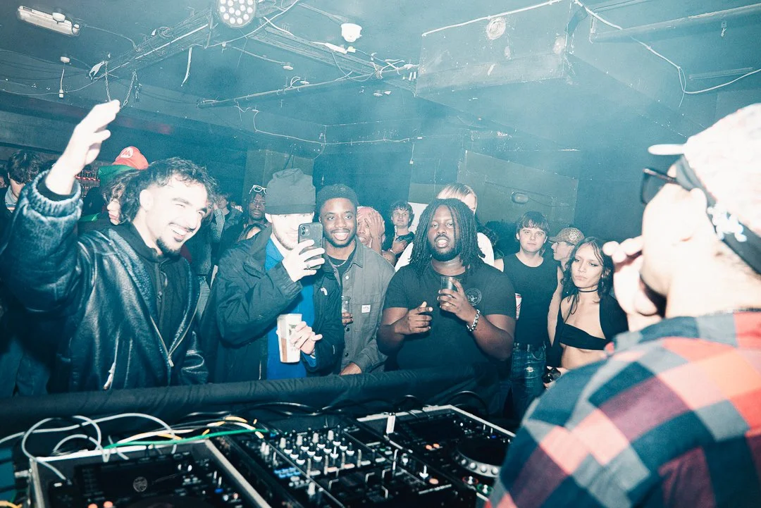People dancing and enjoying music at a nightclub or concert, with DJ equipment in the foreground, in a dimly lit room with blue lighting.