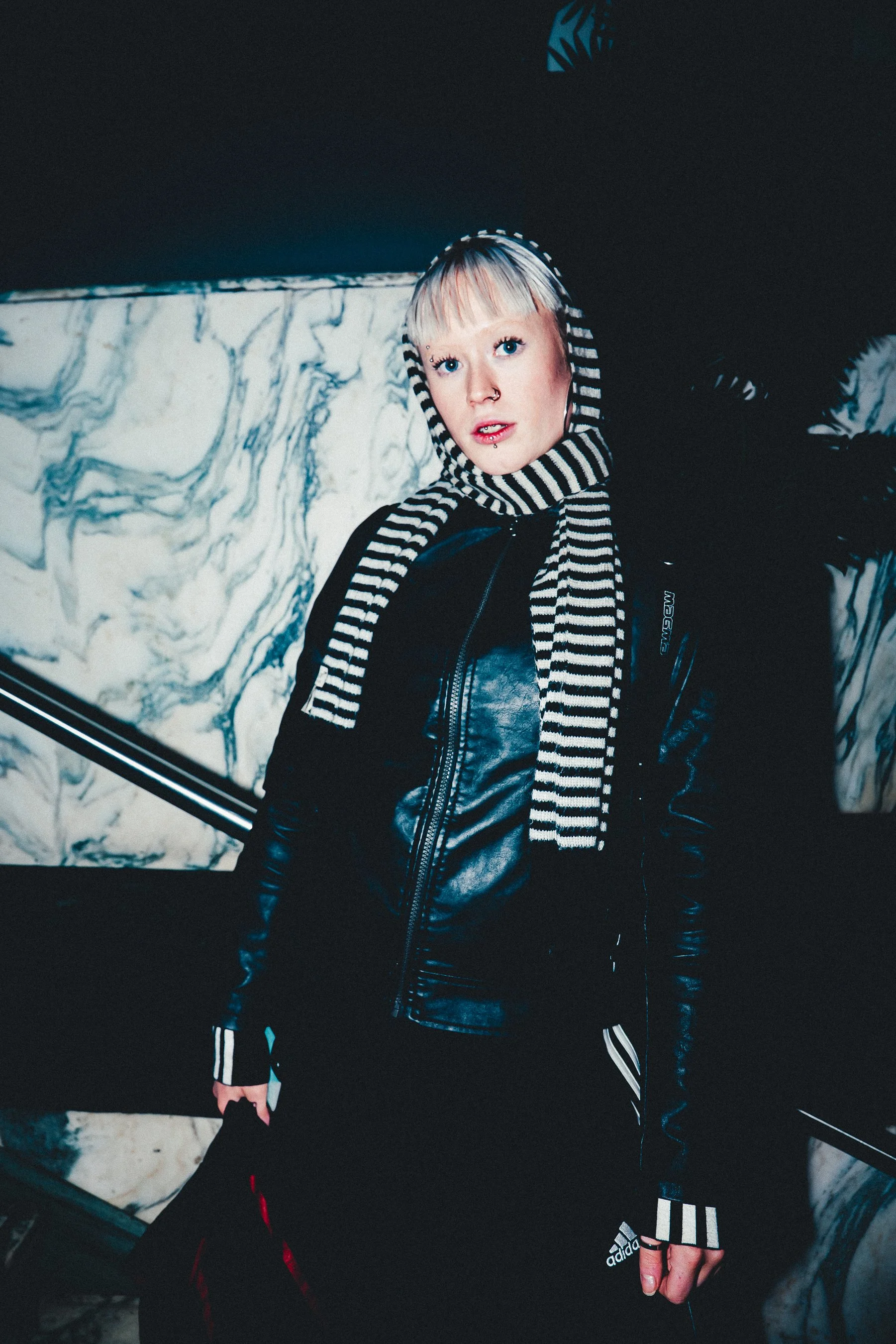 A person with platinum blonde hair, blue eyes, and multiple facial piercings, wearing a black and white striped hooded scarf, black leather jacket, and Adidas sportswear, standing in front of a marble-patterned wall.