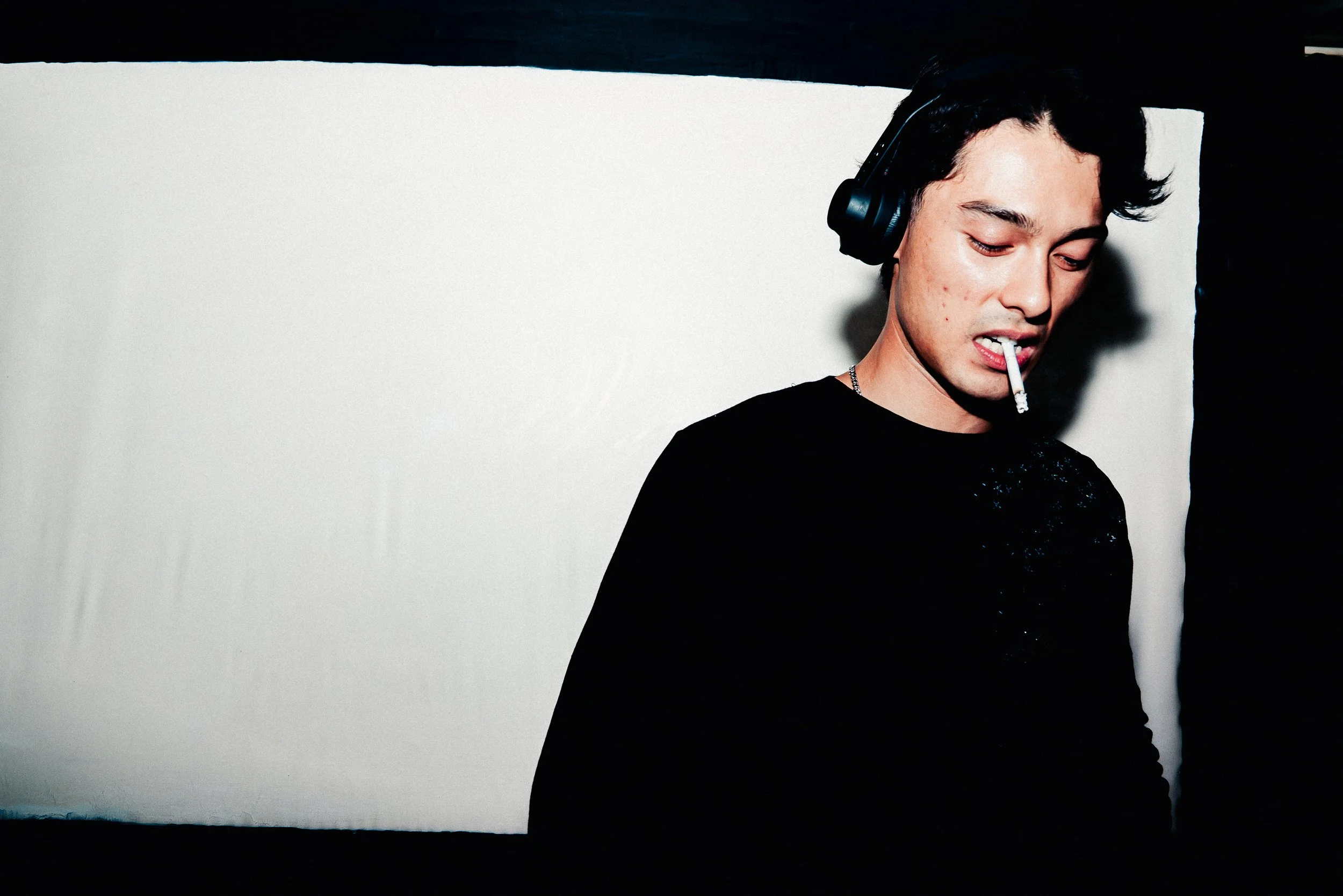 A young man with black hair, wearing headphones and a black shirt, is looking down and biting a cigarette.