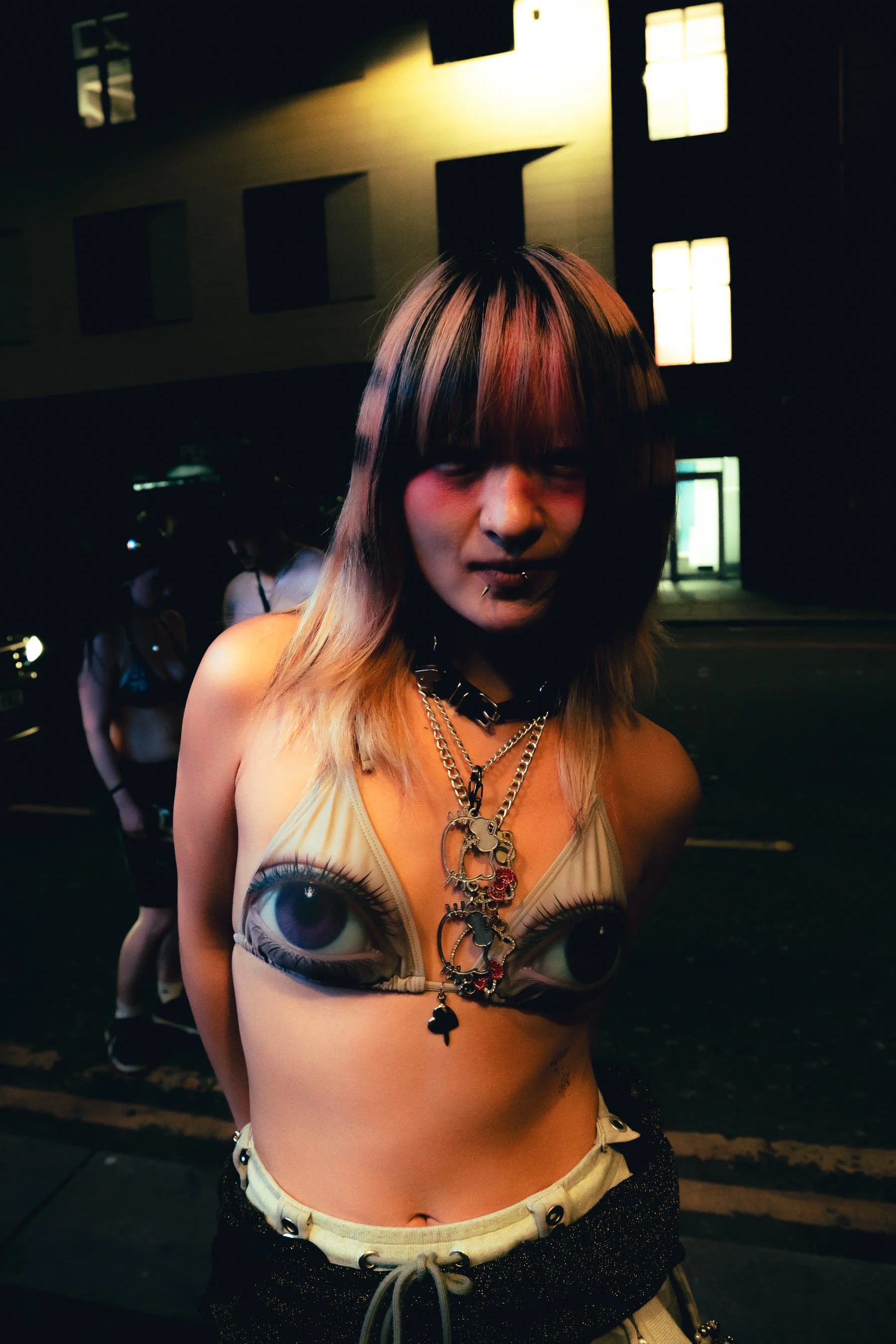 A woman with multi-colored hair stands outside at night, wearing a top with large eye designs and various jewelry, including layered necklaces and a choker.
