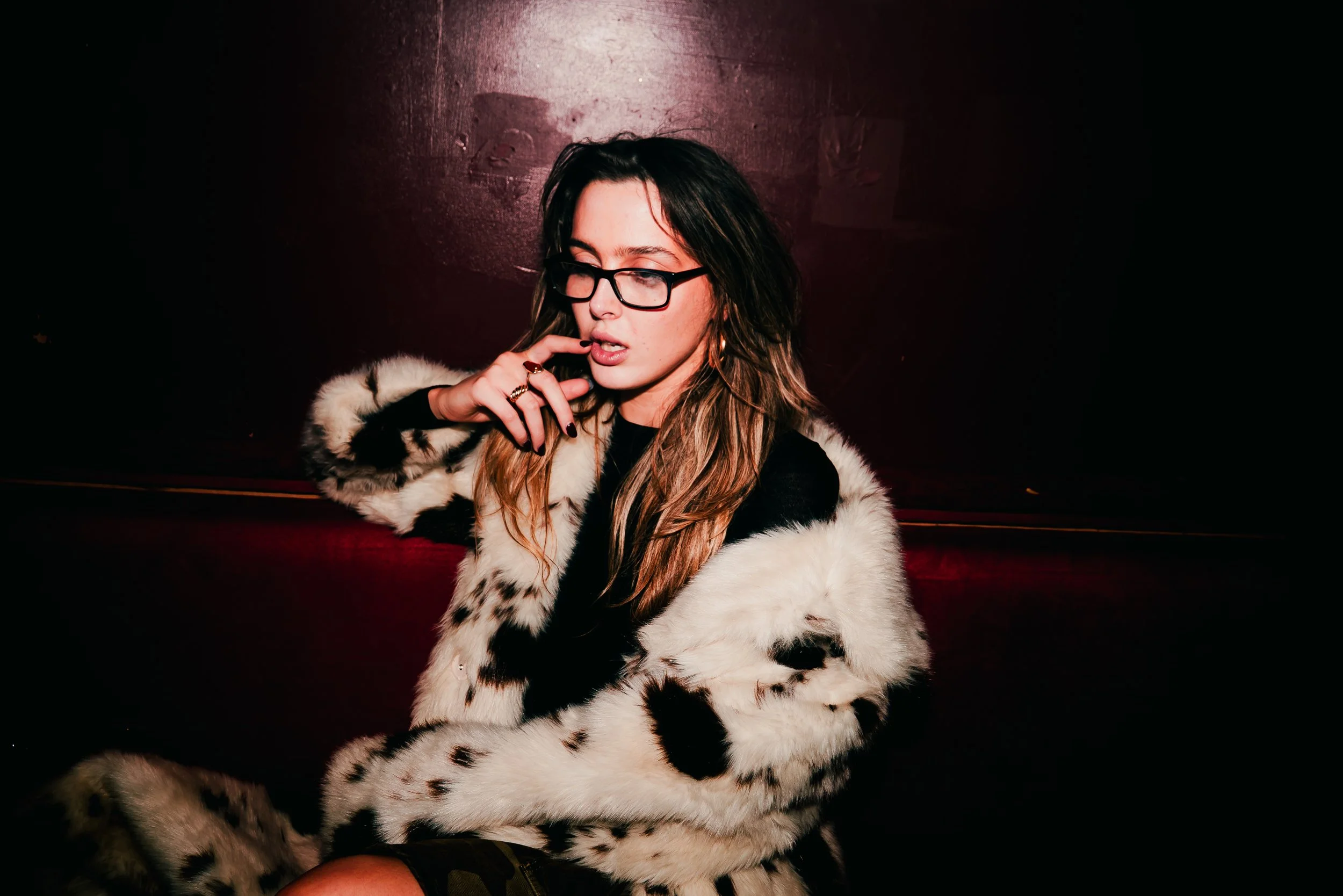 A young woman with glasses wearing a black and white fur coat, sitting against a dark red wall, with her finger near her mouth.