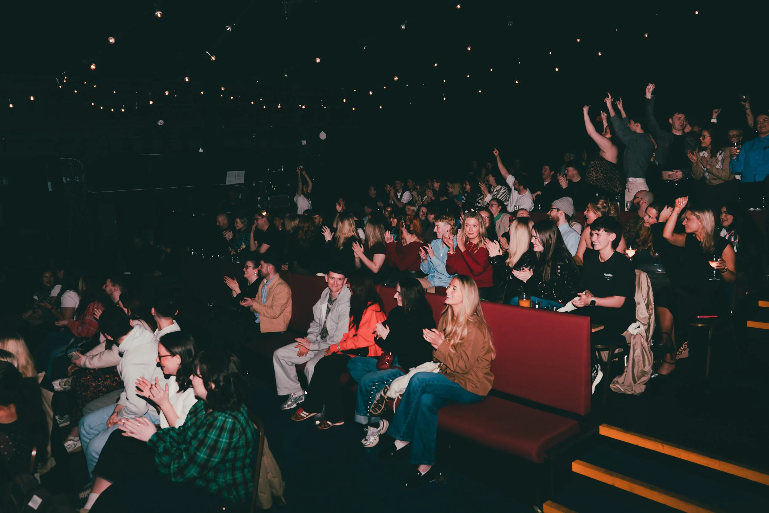Audience at a concert or comedy show in a dark venue, clapping and smiling.