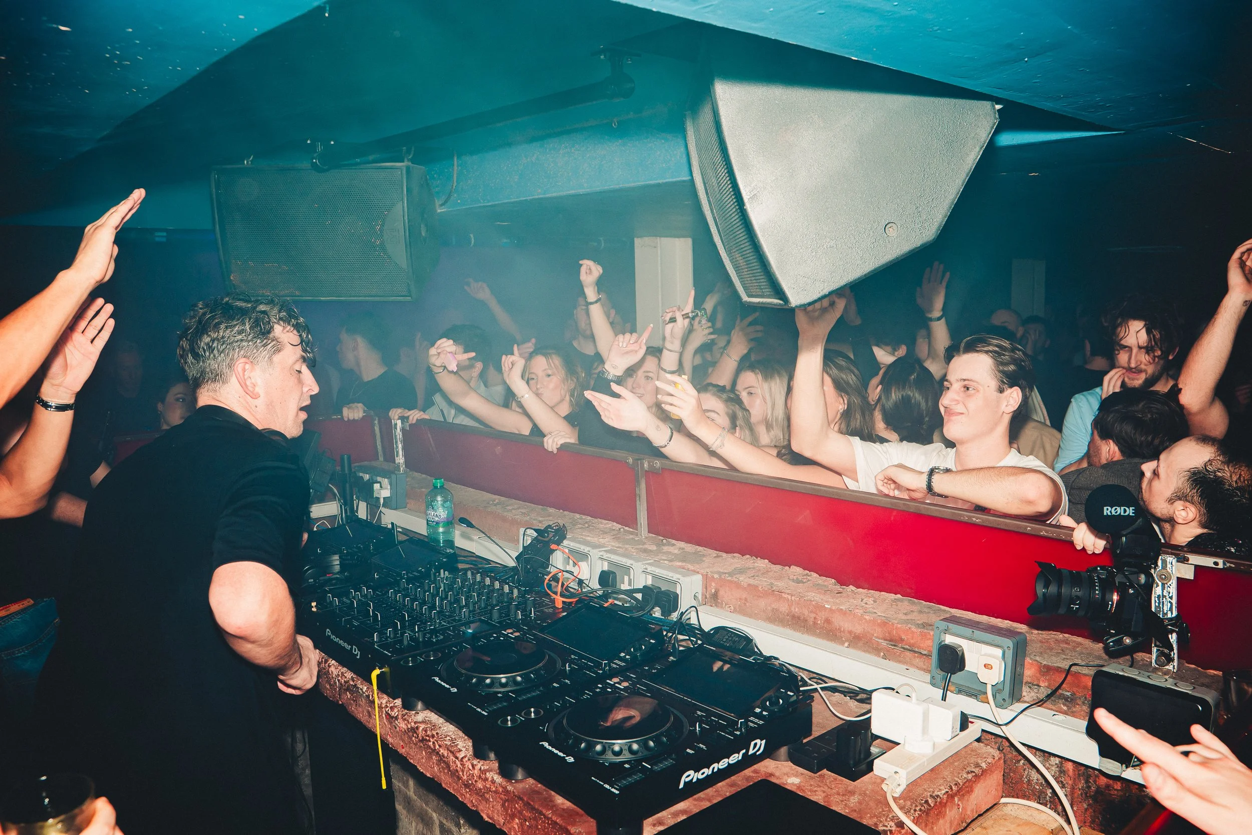 A DJ is performing at a nightclub or event, with a crowd dancing and enjoying the music. The DJ is using sound equipment and there are large speakers overhead. People are raising their hands and celebrating to the music.