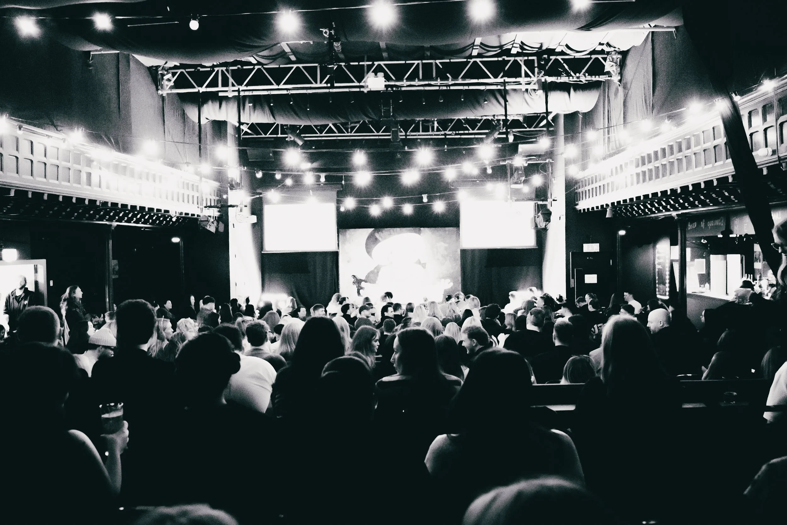 A large crowd of people seated in a theater or event space with a stage at the front, illuminated by multiple bright string lights and spotlights.