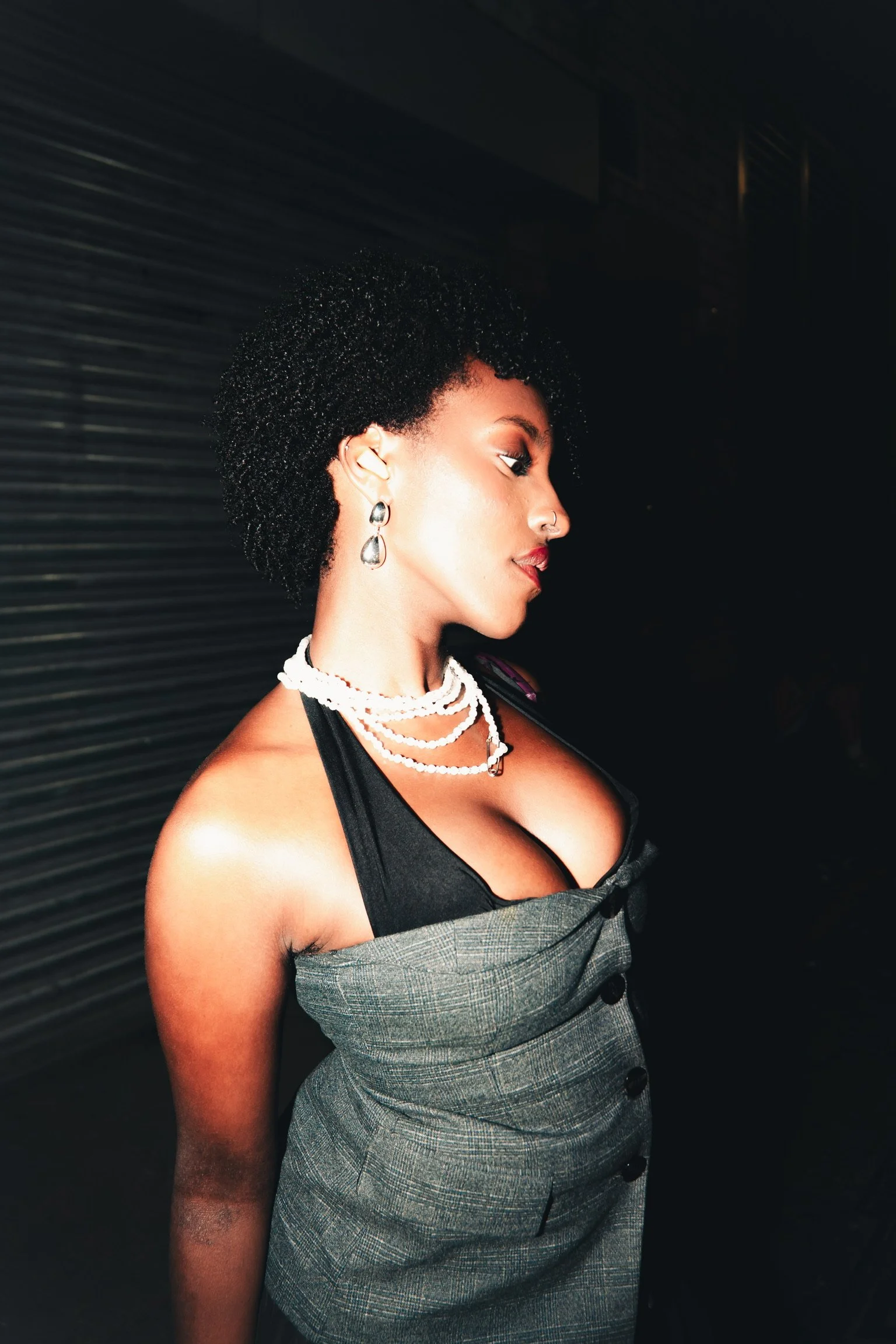 A woman with curly black hair, earrings, and layered necklaces wearing a black top and plaid skirt in front of a dark background.