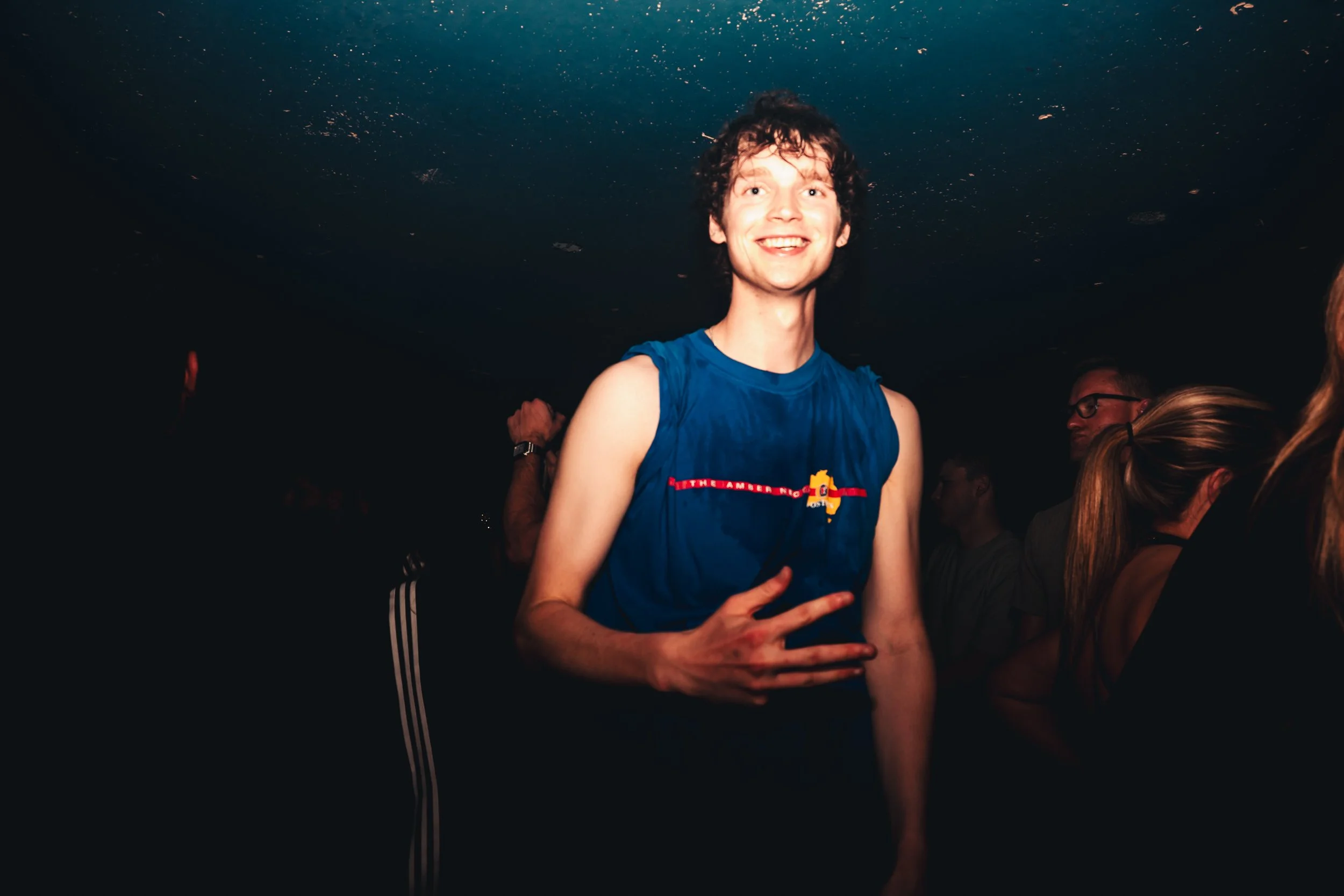 A young man with curly hair and a big smile, wearing a blue sleeveless shirt, is posing with a peace sign gesture, standing in a dimly lit space with a crowd around him.