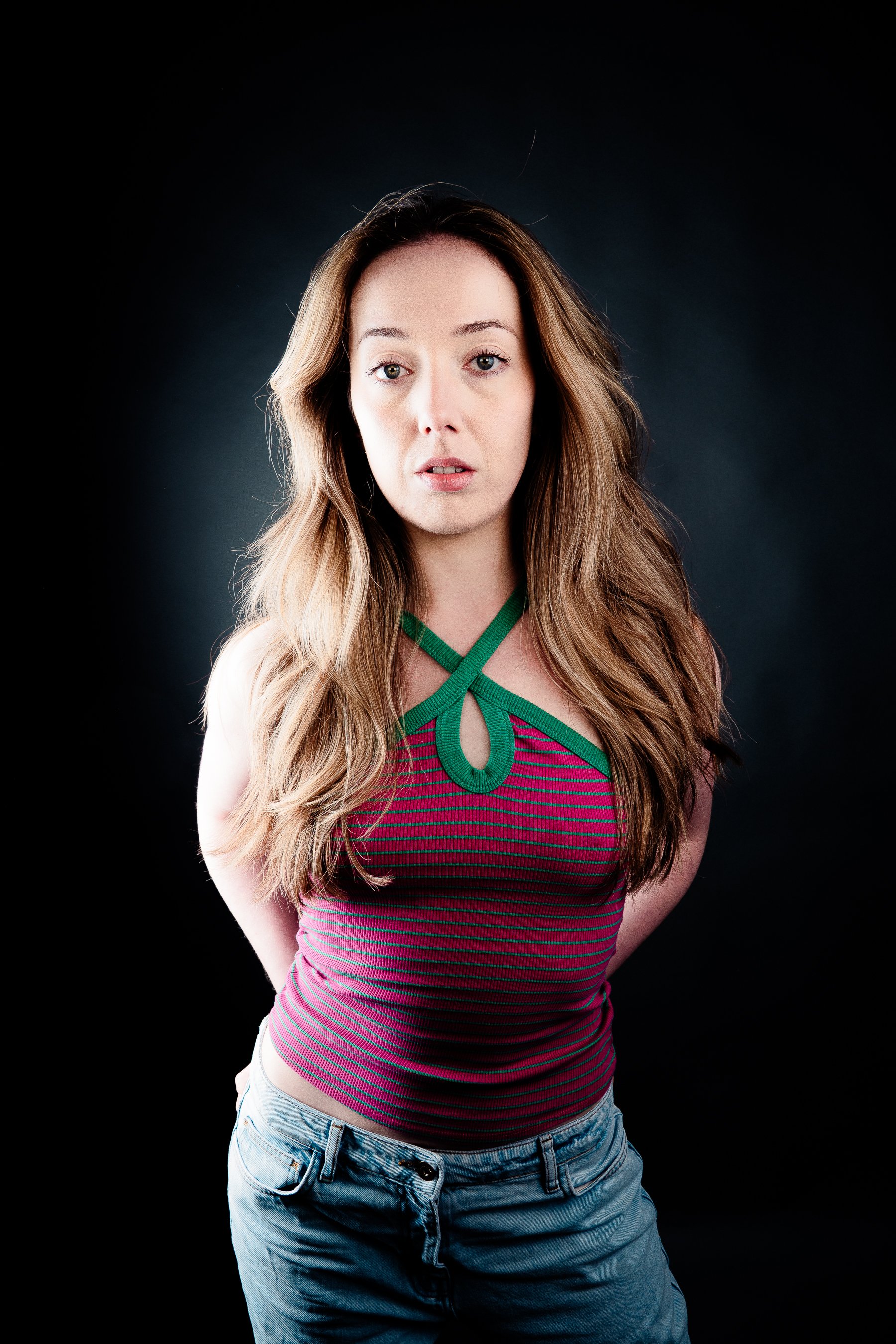 A young woman with long wavy blonde hair and light skin poses against a dark background, wearing a striped pink and green top with a keyhole detail and light blue denim shorts.