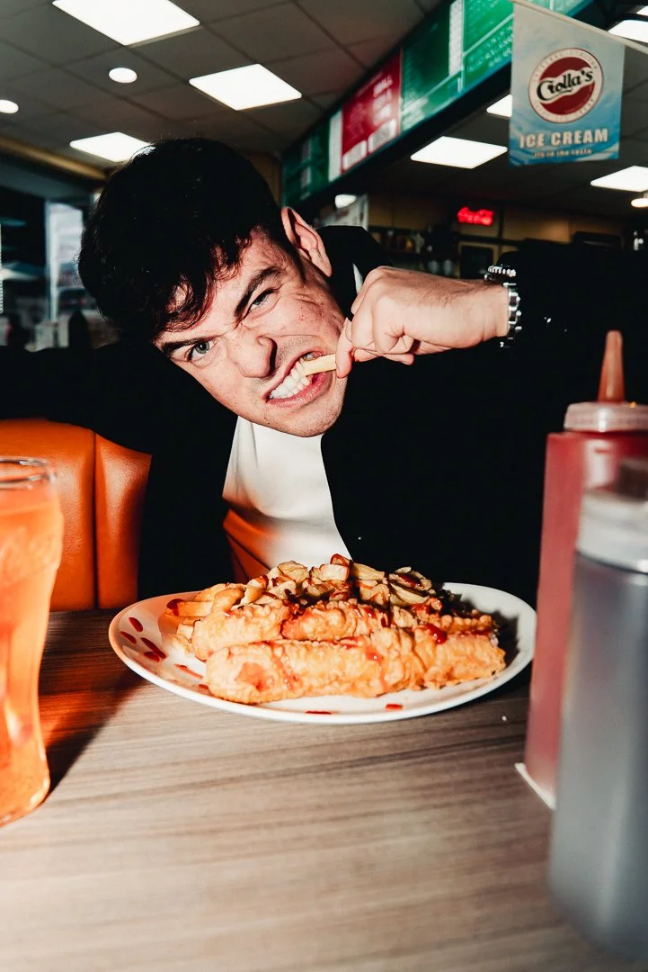 Man sitting at a diner table, about to eat a large slice of pie, making a playful angry face, with ketchup and mustard bottles on the table and a fast food restaurant interior in the background.