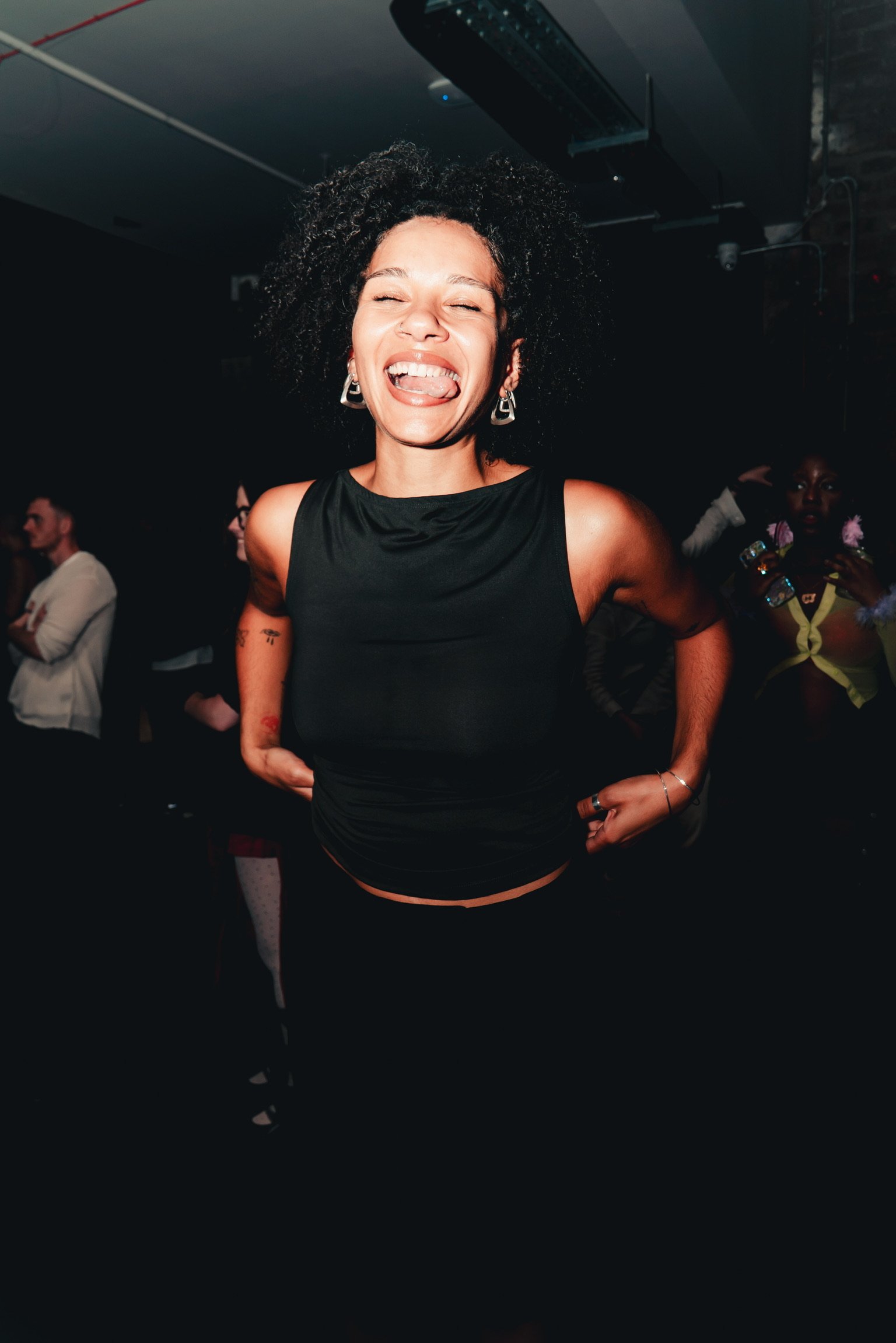 A woman smiling with her eyes closed and sticking out her tongue, wearing a black sleeveless top and earrings, standing in a dark environment with other people in the background.