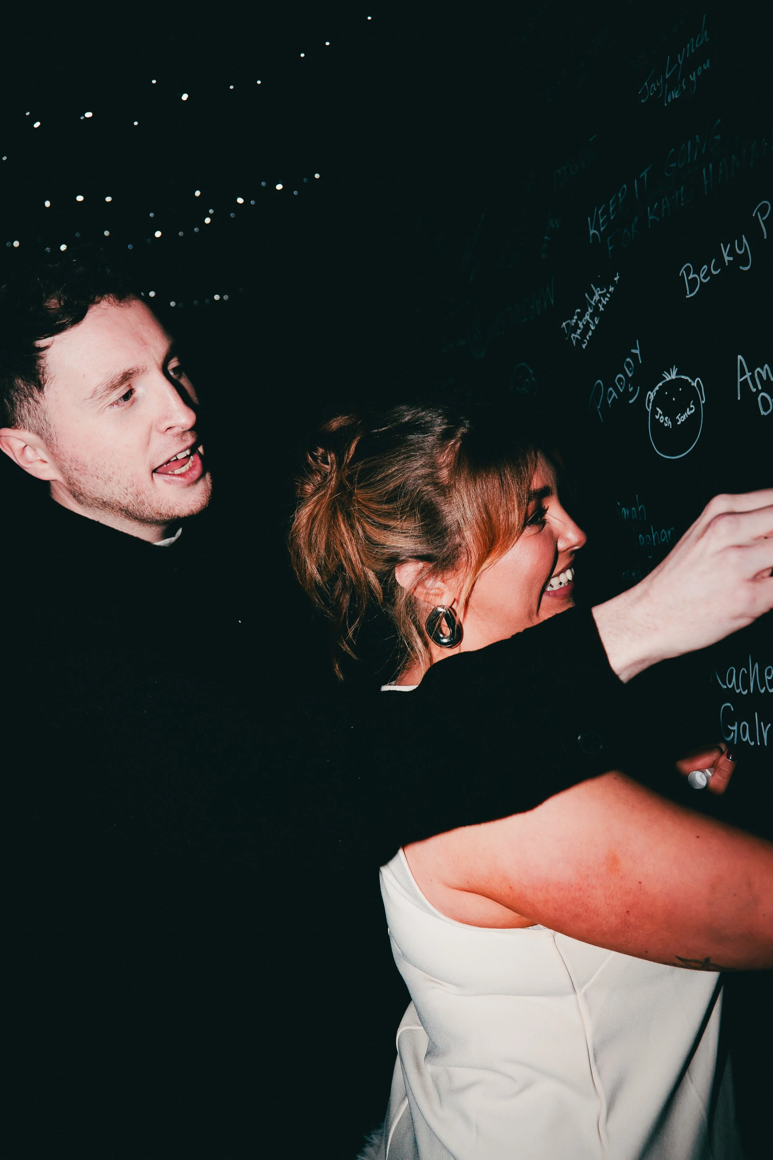 A man and woman smiling and writing on a black chalkboard wall decorated with string lights.