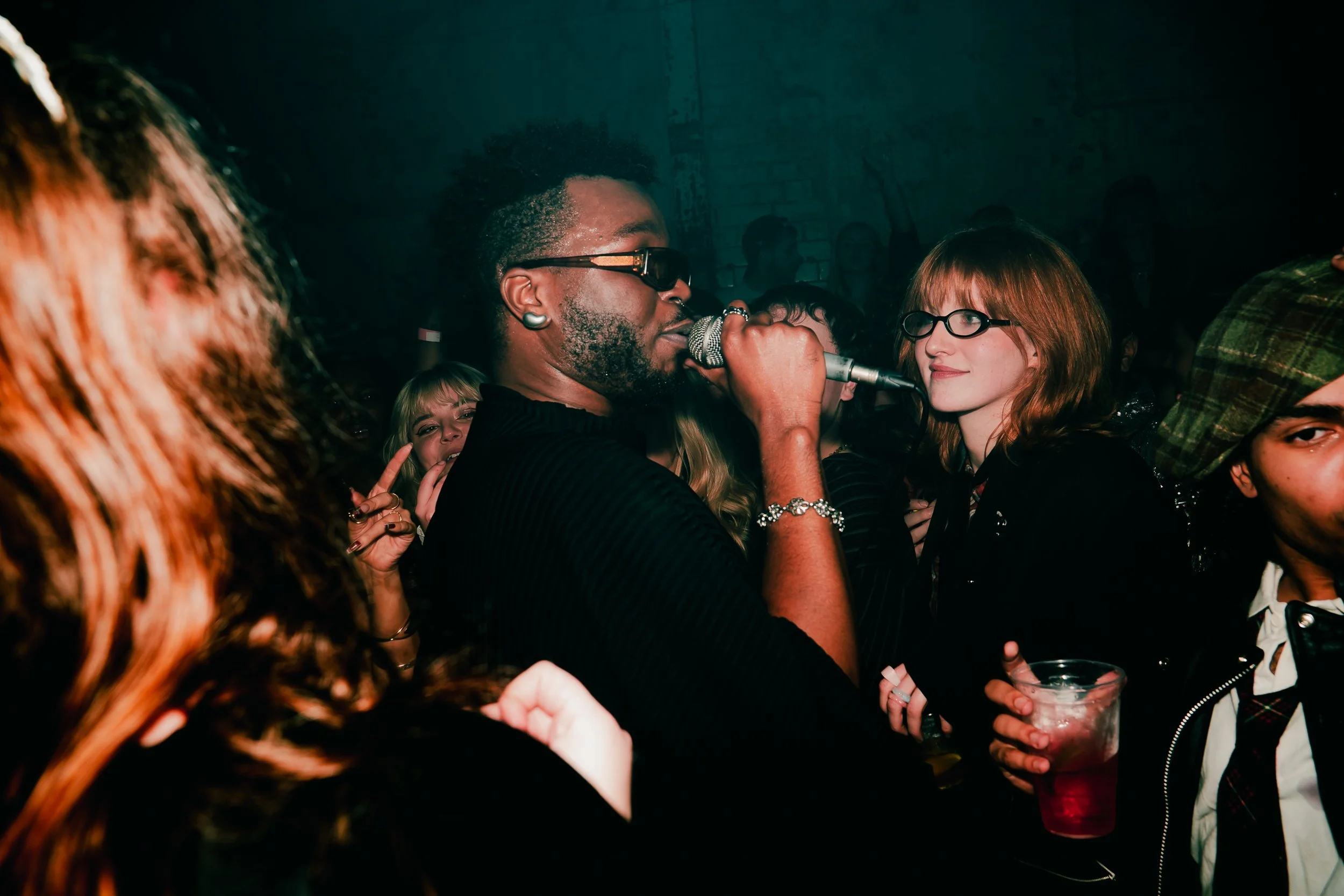 A man with sunglasses singing into a microphone at a party surrounded by women, some holding drinks and smiling.
