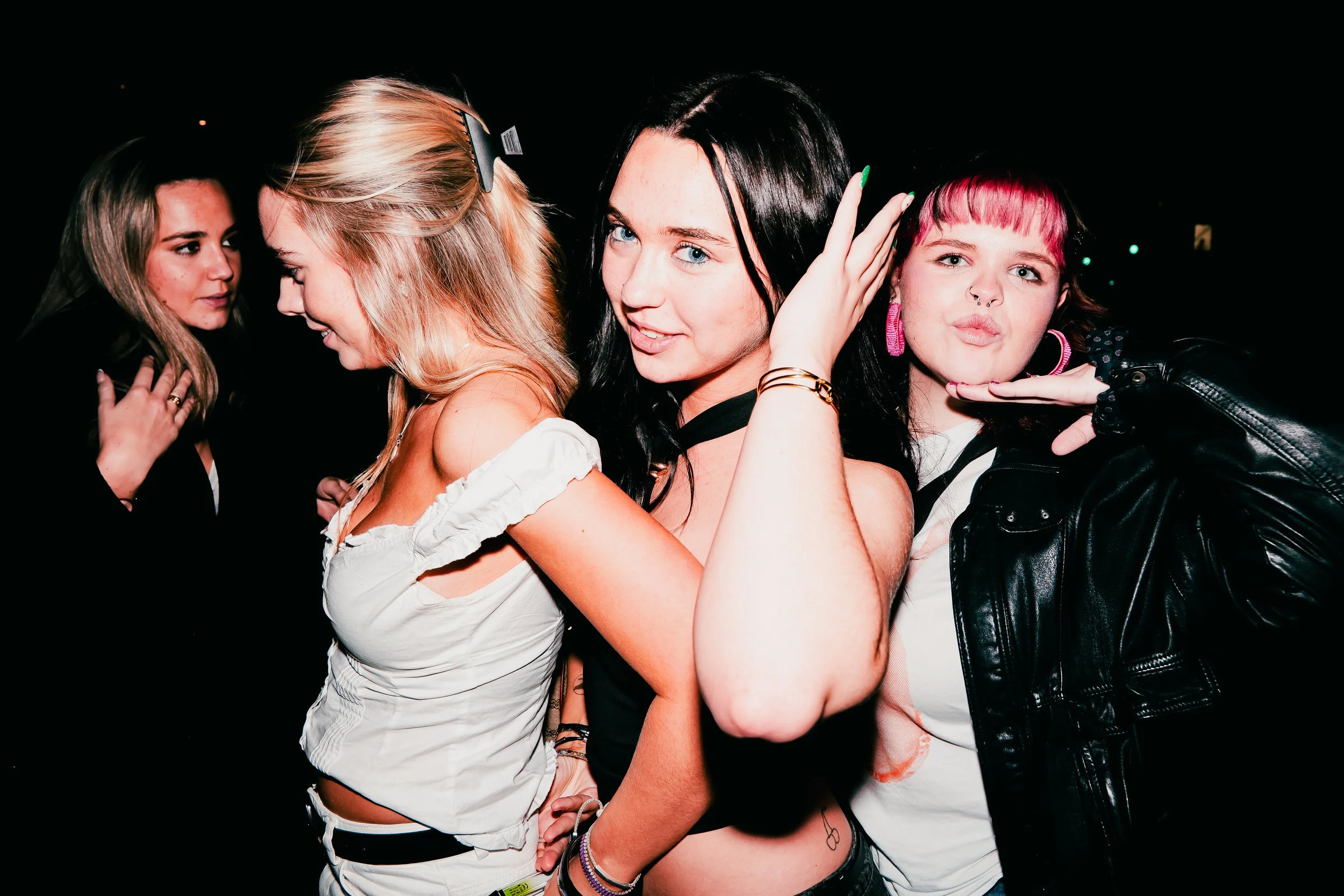 Group of young women at a party or club, dressed casually, with one woman in the center adjusting her hair and the others either engaged in conversation or looking at each other, dark background.