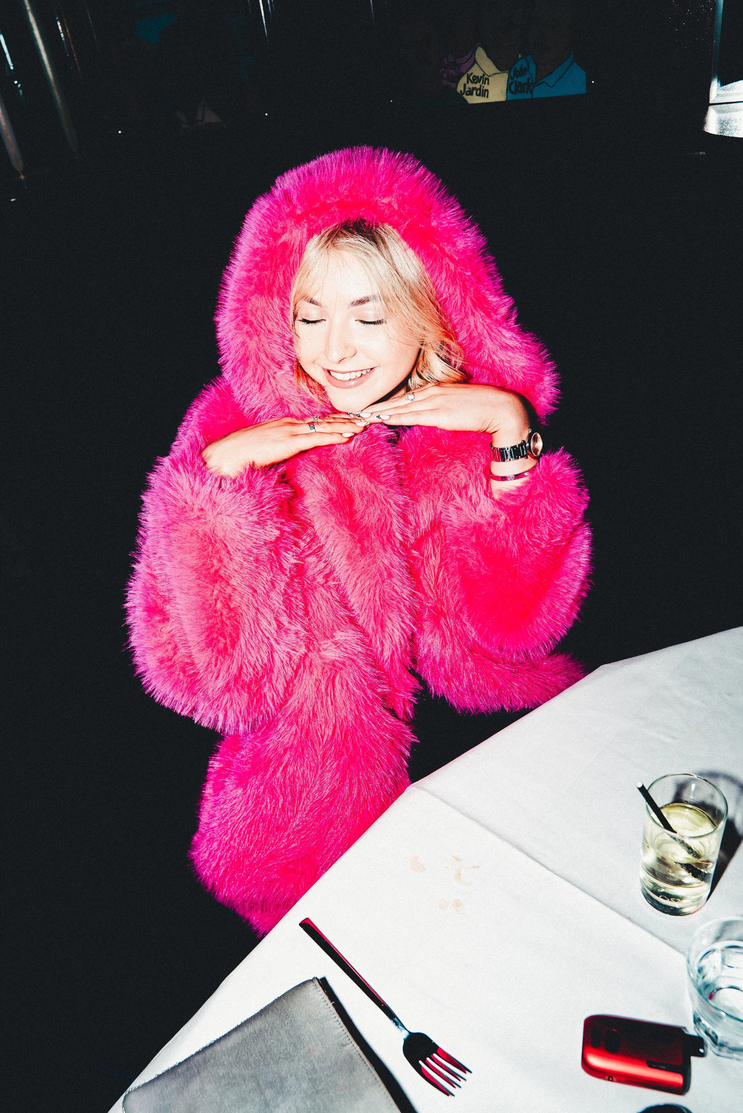 A woman dressed in a bright pink fur coat with a hood is sitting at a table with her eyes closed and hands under her chin, smiling. There are drinks and a phone on the table.