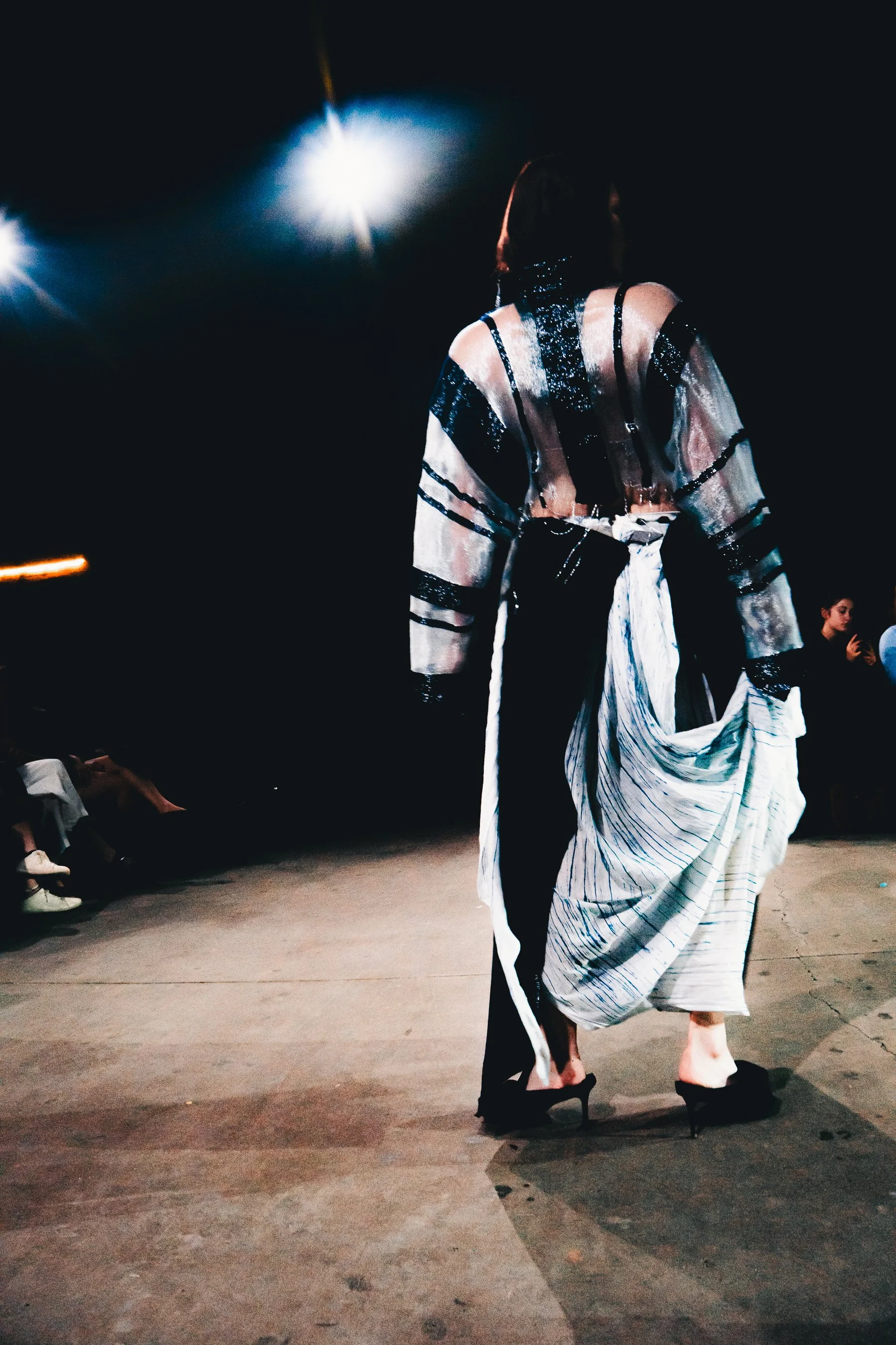 A fashion model walking on a runway stage, wearing high heels and an artistic, layered outfit, with spotlights illuminating her against a dark background.